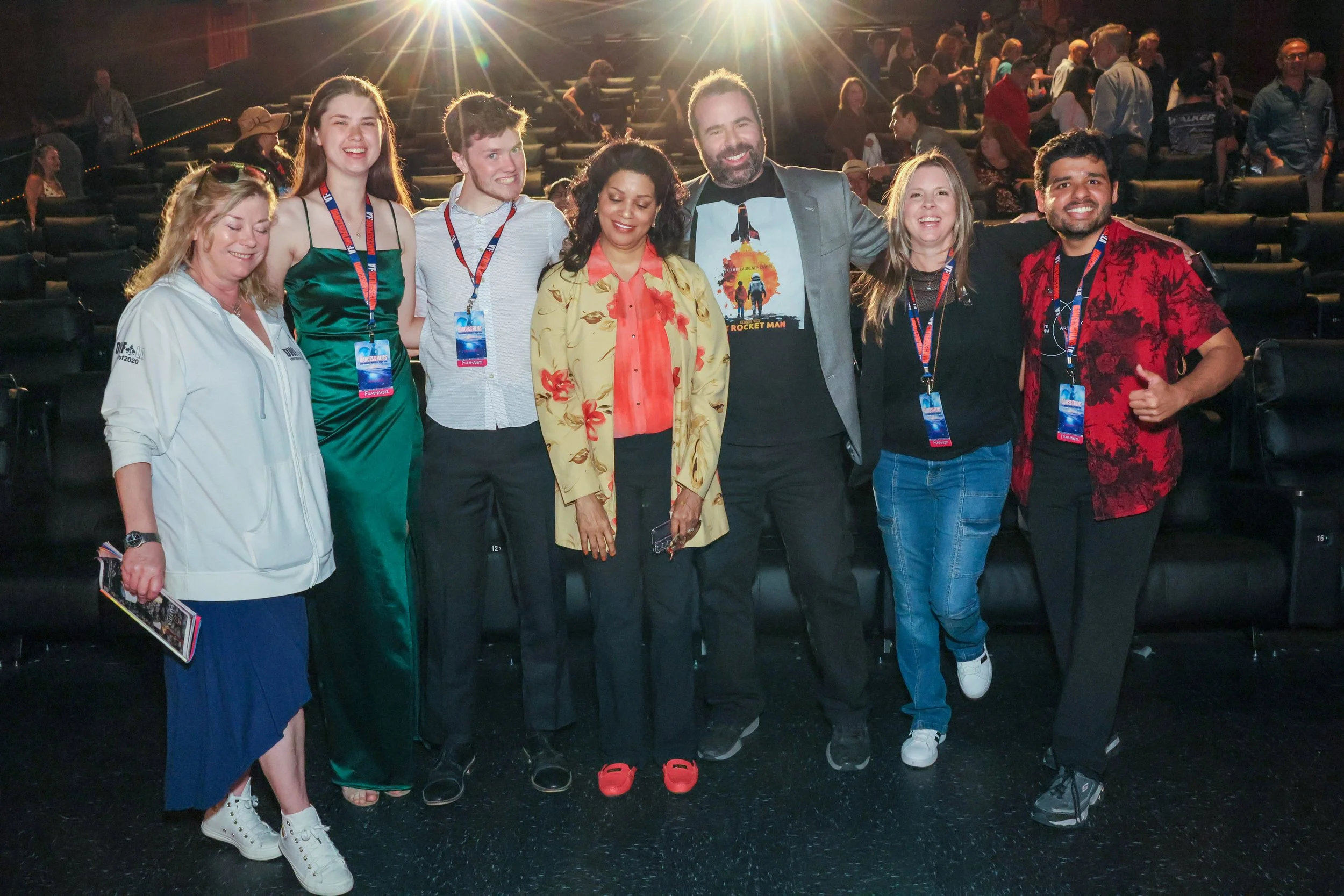 Group of people standing together in a theater or conference hall with bright light behind them, smiling for the camera.