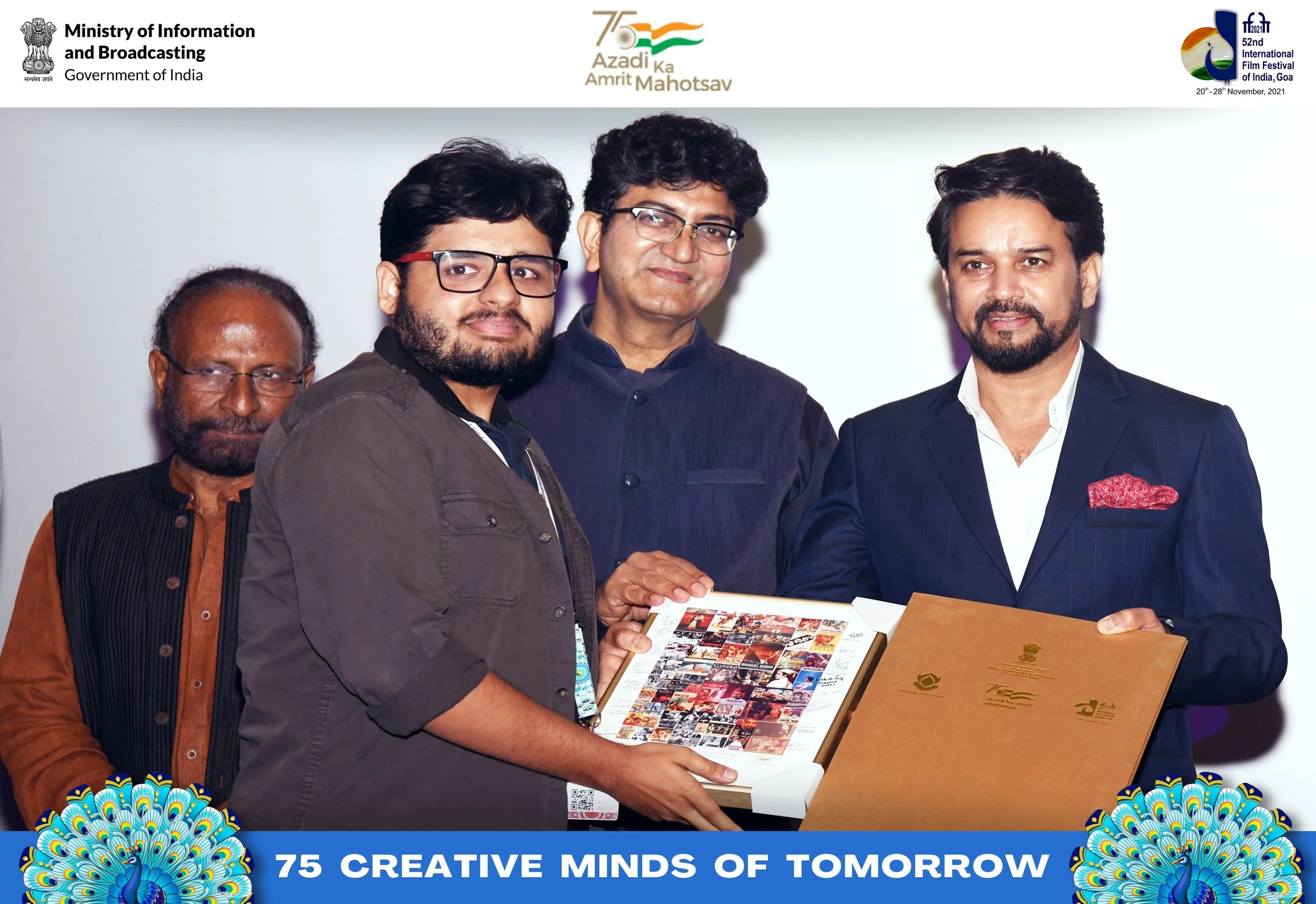 Four men at an event, with one man holding a book and a certificate or folder, standing in front of a white background with logos of the Indian government and film festival, and a banner reading '75 Creative Minds of Tomorrow' with peacock graphics.