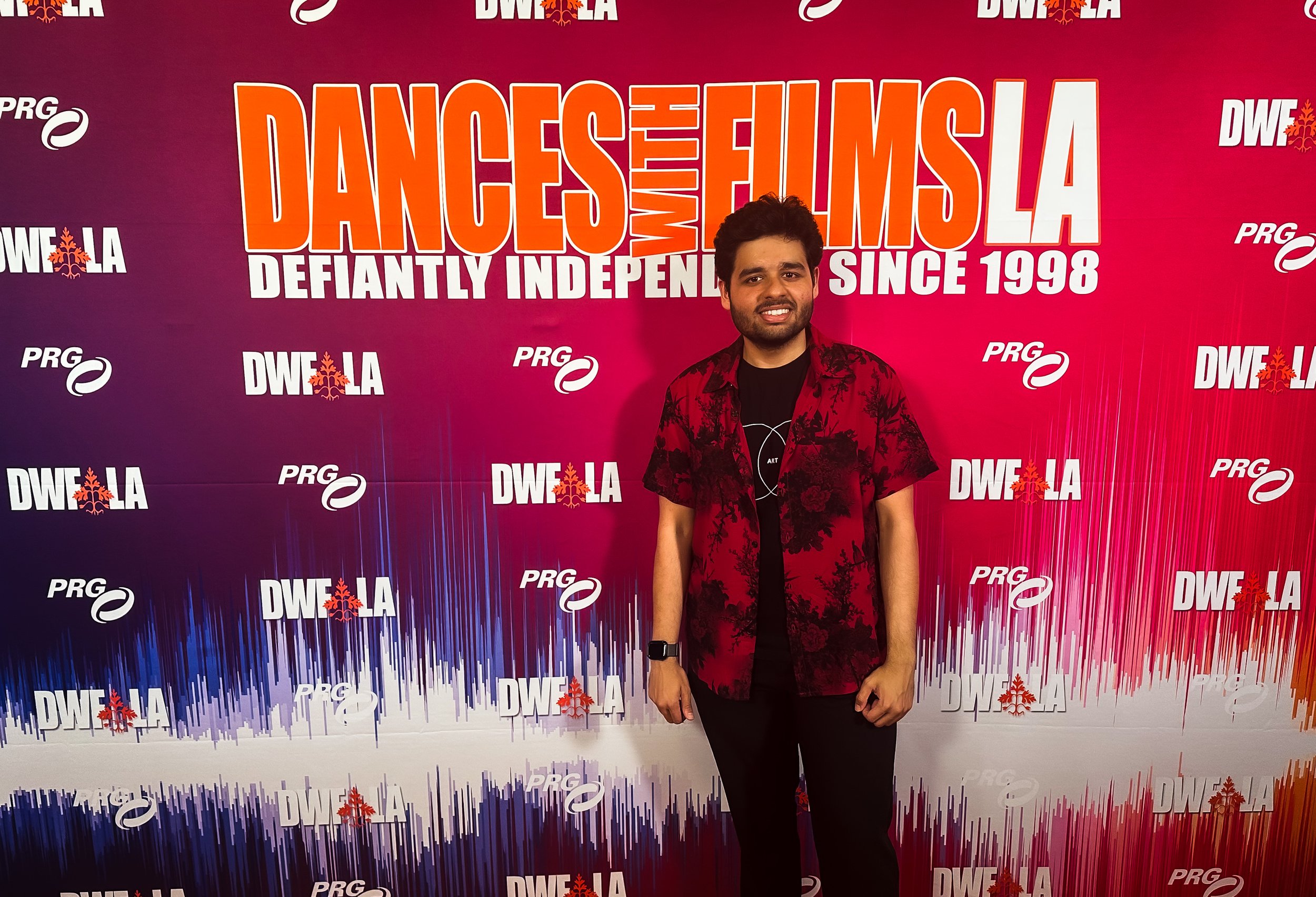 A man standing in front of a pink and purple backdrop with the text 'DANCES WITH FILMS LA DEFINITELY INDEPENDENT SINCE 1998' and logos, wearing a black shirt, red floral short-sleeve shirt, black pants, and a smartwatch, smiling at the camera.
