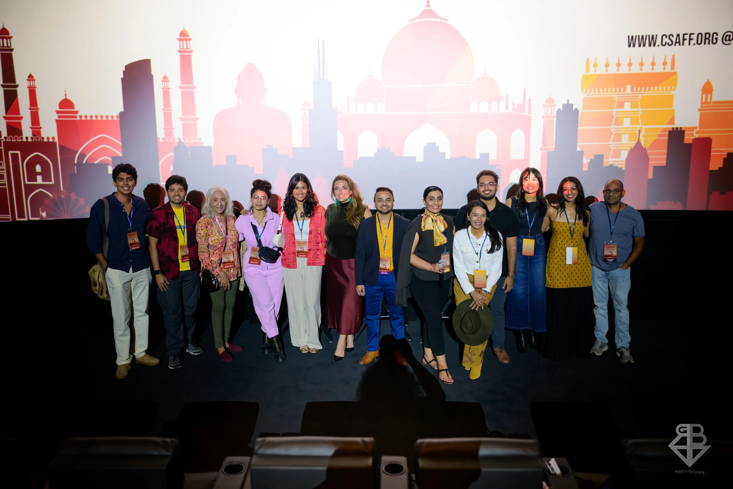 Group of diverse people standing on a stage in front of a large screen displaying an artistic silhouette of Indian landmarks, with some wearing conference badges.