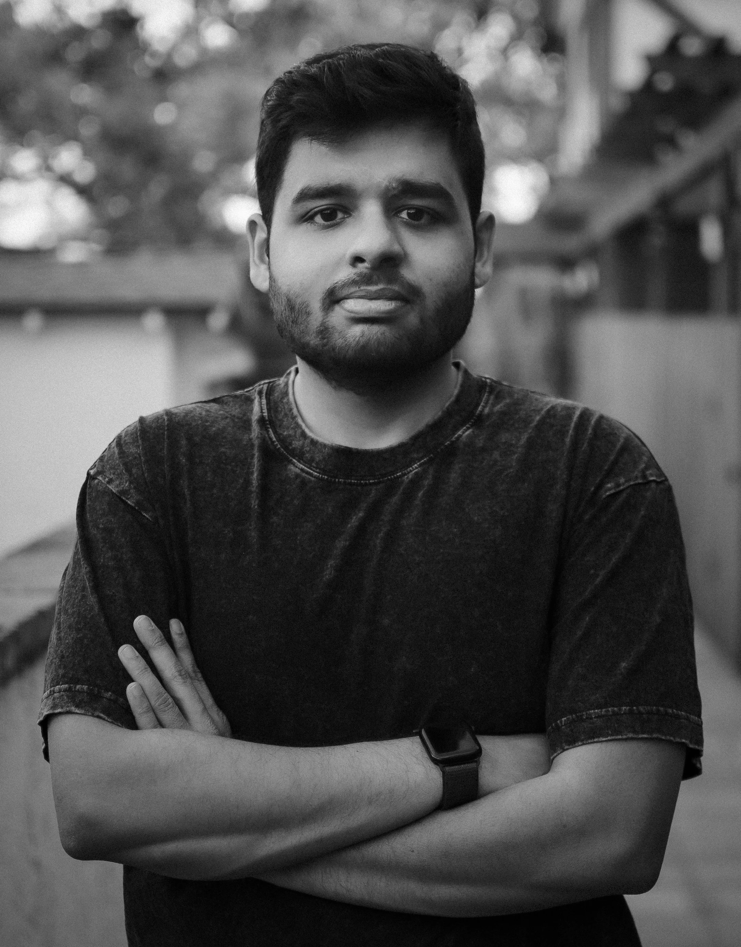 A young man with dark hair and a beard, standing outdoors with his arms crossed, wearing a dark T-shirt and a smartwatch, in a blurred background with trees and buildings.