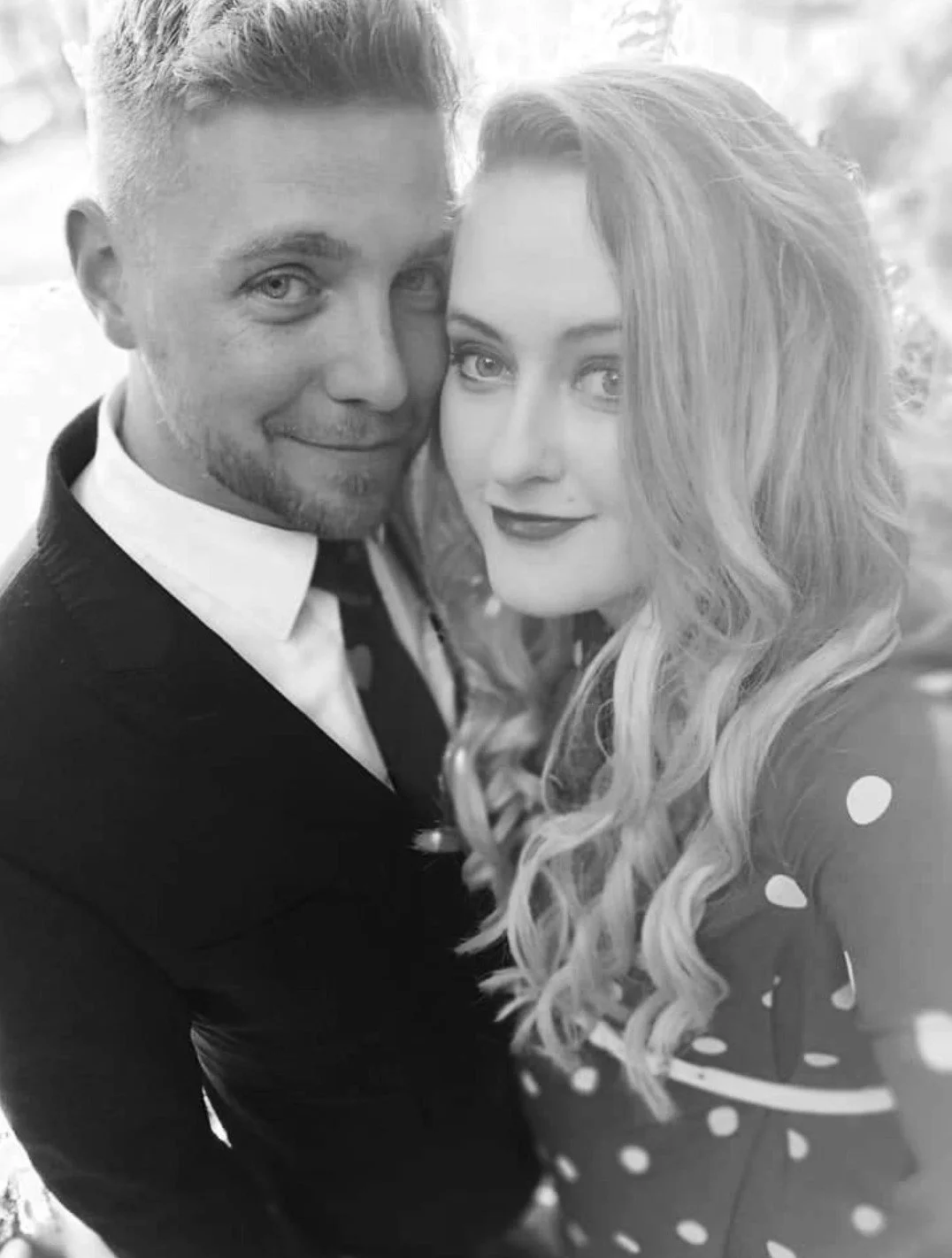 A black-and-white photo of a man and woman close together, smiling at the camera. The man has short hair, light facial hair, and is dressed in a suit. The woman has long, wavy hair and is wearing a polka-dot top.