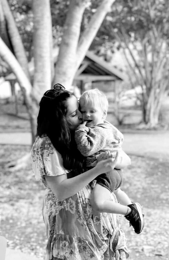 A woman holding a young boy in an outdoor park setting, sharing a kiss on the cheek, with trees in the background.