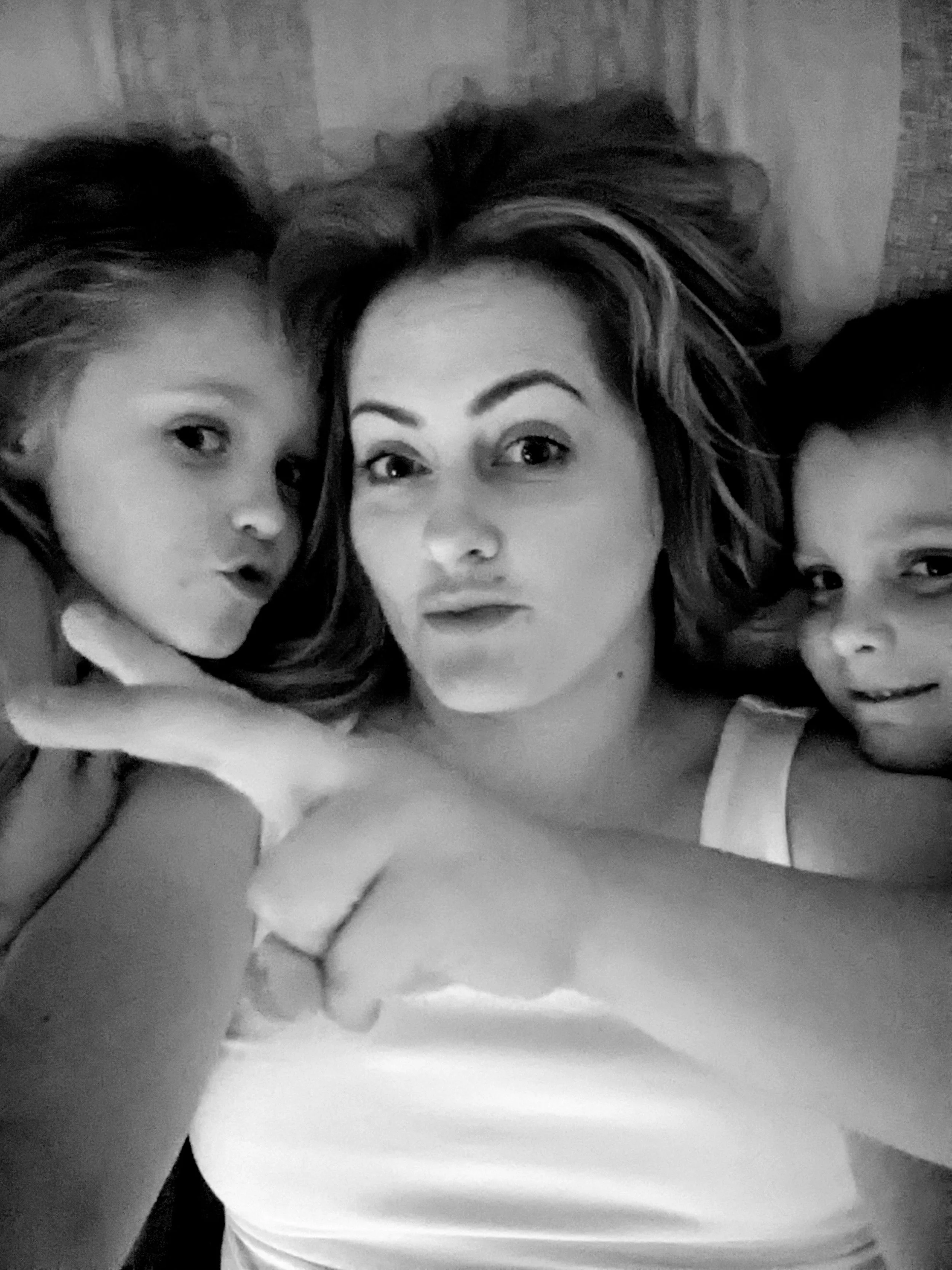 Black-and-white selfie of three women lying on a bed with their heads close together, taking a photo with a smartphone.