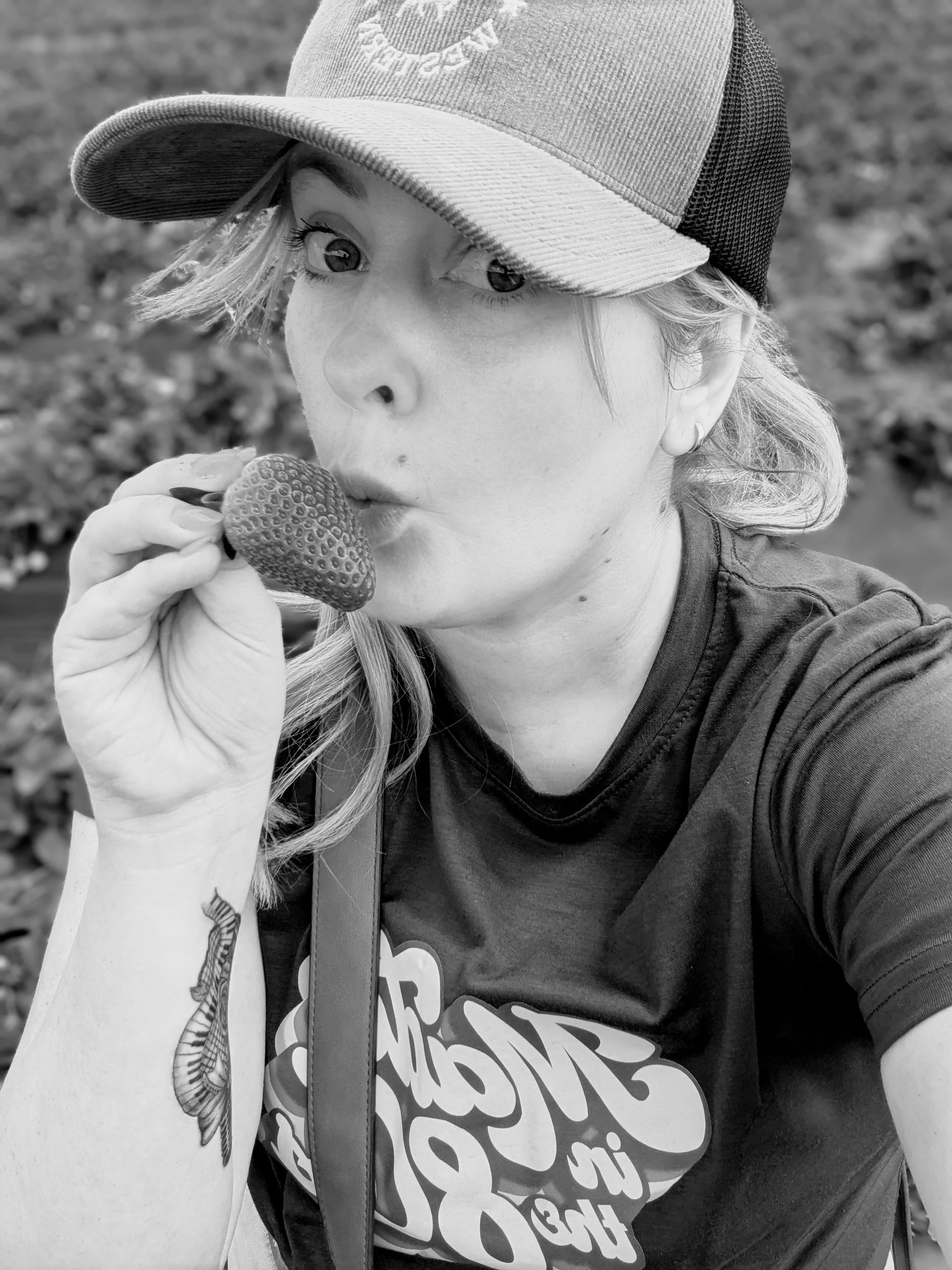 A woman wearing a cap and t-shirt holding and biting a strawberry in an outdoor setting.