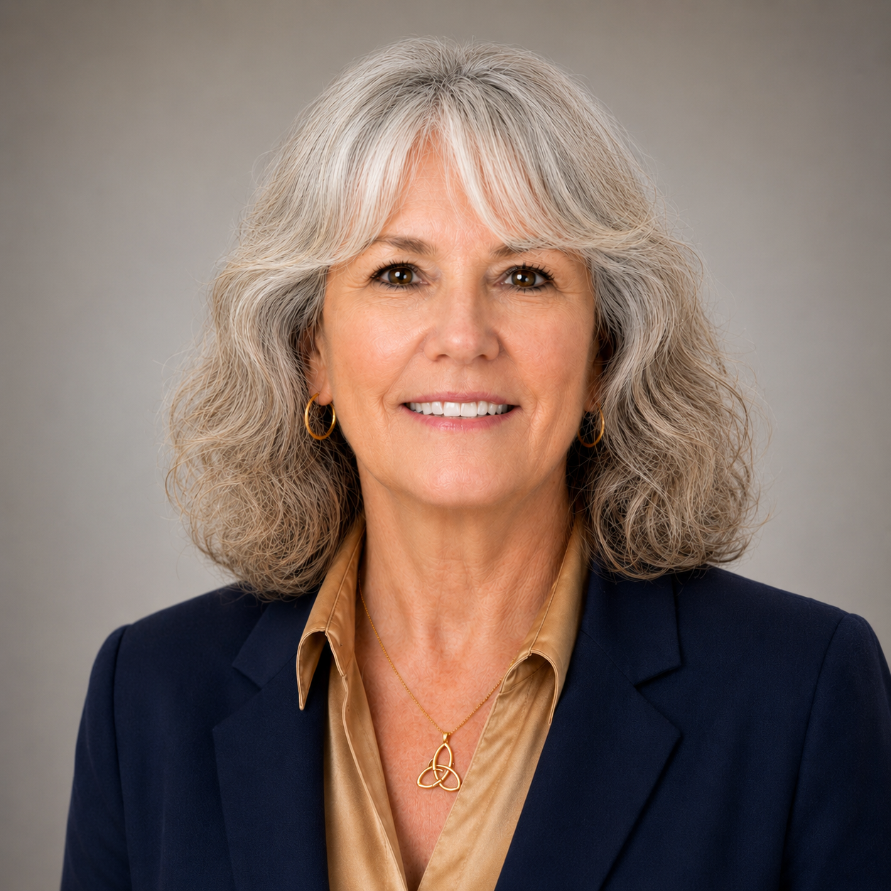 A portrait of a smiling older woman with curly gray hair, wearing a navy blazer, gold jewelry, and a tan blouse, against a neutral gray background.