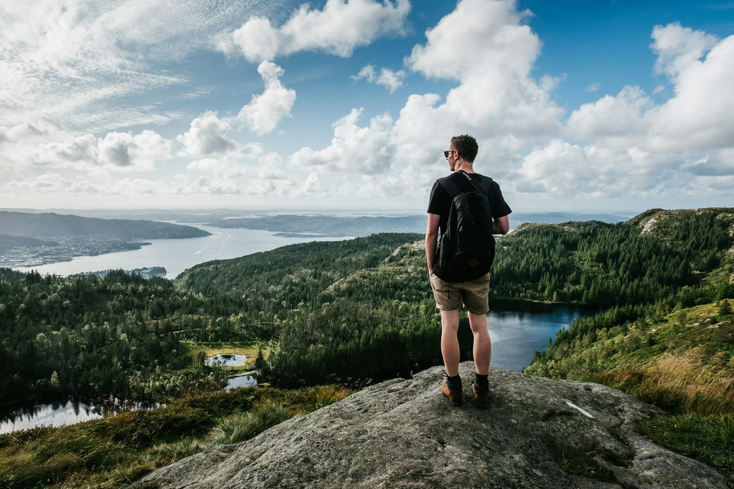 A man with a backpack standing on a large rock, overlooking a scenic landscape of lush green hills, lakes, and a river winding through the valley under a partly cloudy blue sky.