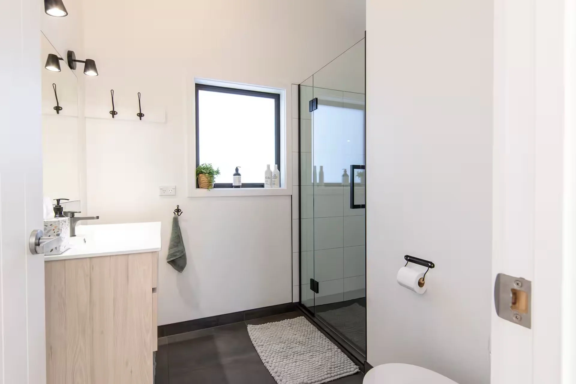 Modern bathroom with white walls, black floor tiles, and a glass shower enclosure. There is a small vanity with a sink, a window with a plant and bottles nearby, and a towel hanging on a hook. The bathroom also features a toilet paper holder and a bath mat.