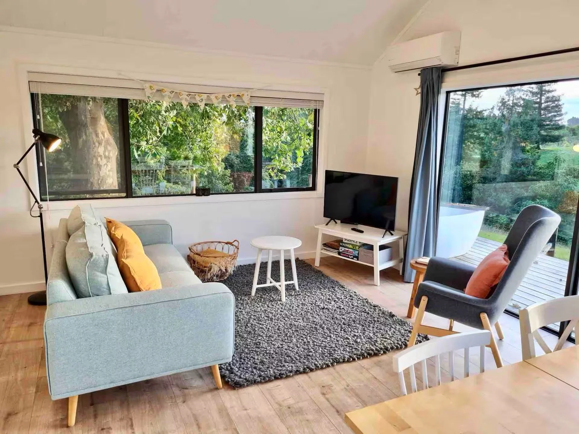 Living room with light blue sofa with yellow pillows, a black floor lamp, a white small round table, a black rug, a white TV stand with a flat-screen TV, and large windows showing green trees outside, with some curtains and an air conditioner unit on the wall.