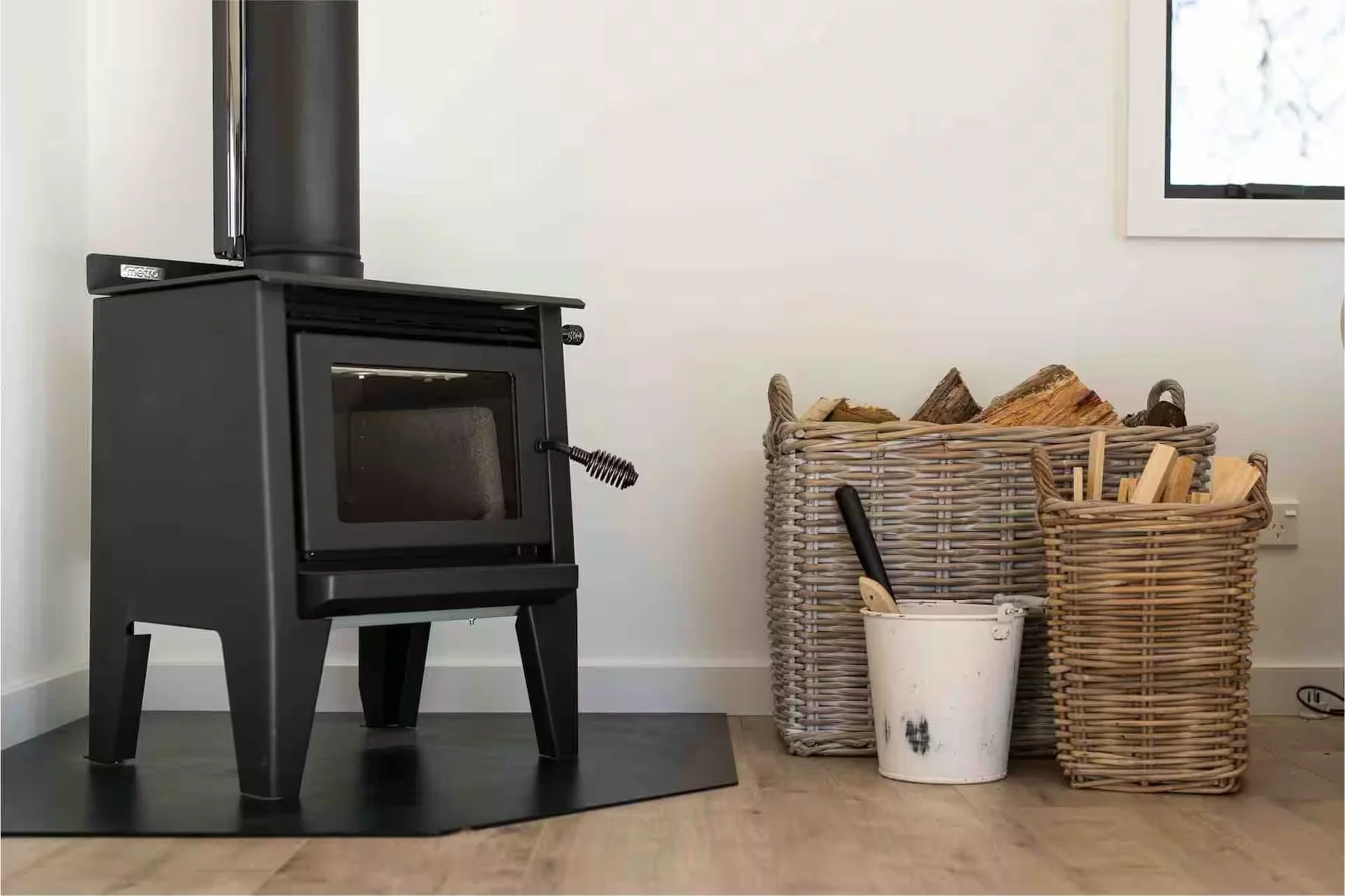 A black wood stove in a room with woven baskets filled with firewood, a white bucket with black marks, and part of a window showing outside trees.