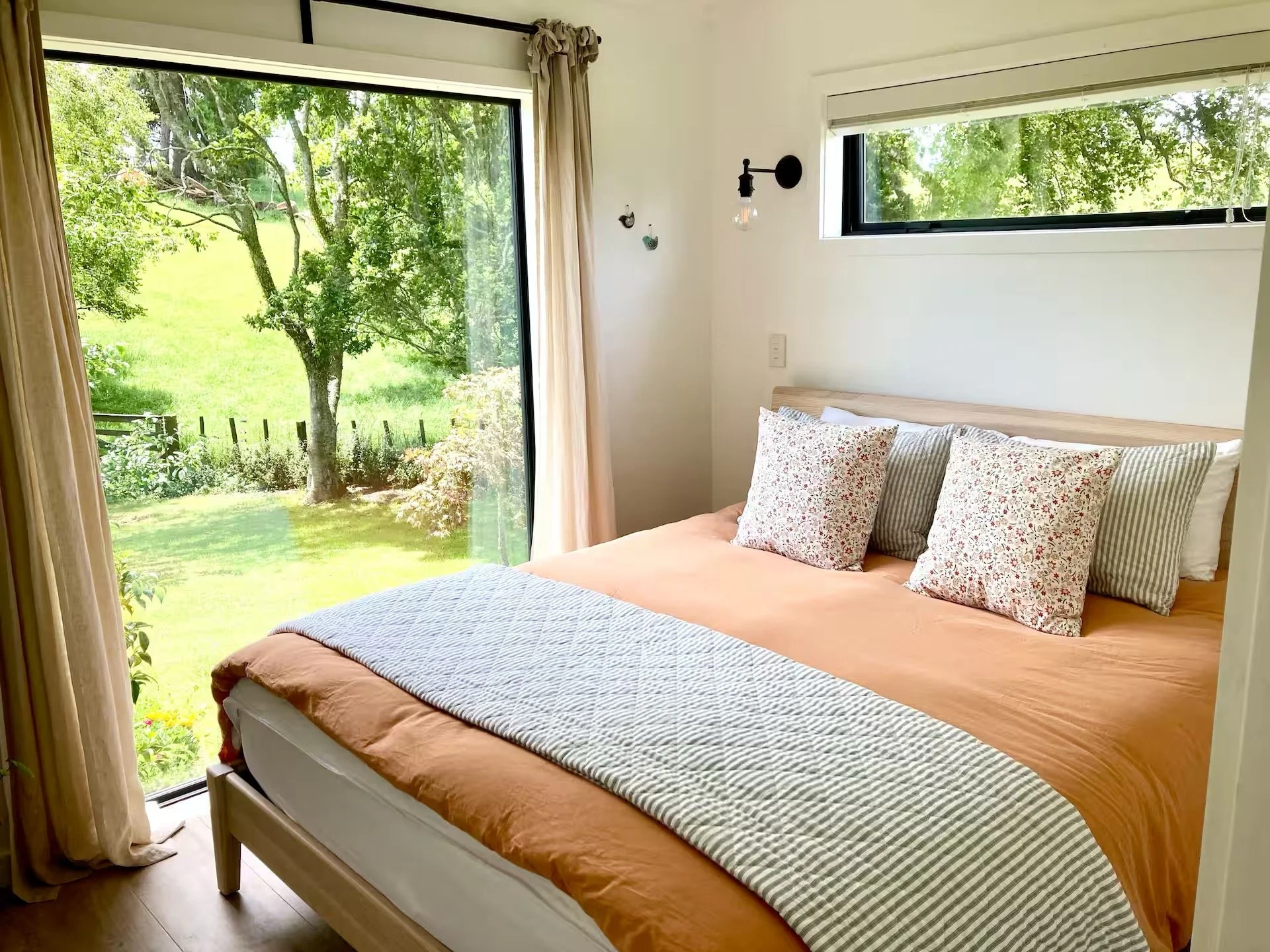 A bedroom with a large sliding glass door opening to a green garden with trees. The bed has orange bedding, a striped blanket at the foot, and multiple patterned pillows. There is a horizontal window above the bed, a wall-mounted black light fixture, and wall hooks.