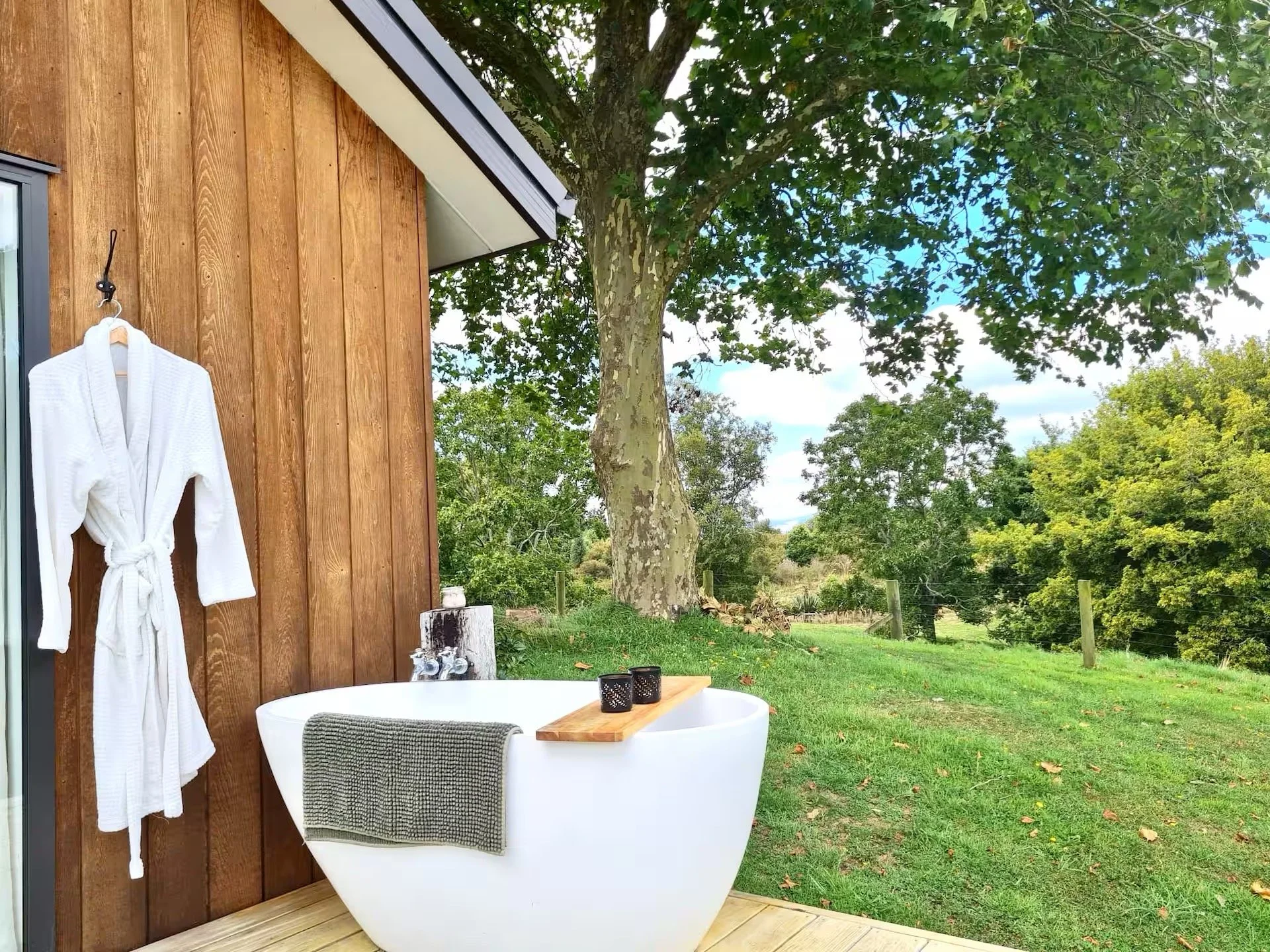 An outdoor bathtub next to a wooden wall with a white bathrobe hanging on a hook, overlooking lush green trees and a grassy area.