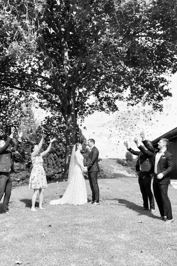 Black and white photo of a wedding ceremony outdoors under a large tree with a bride and groom holding hands, surrounded by friends throwing flower petals and celebrating.