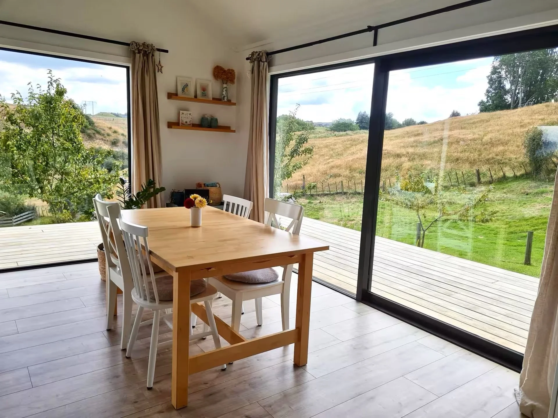 A dining area with a wooden table and four white chairs, decorated with a small vase of flowers. The room has large glass sliding doors that open to a wooden deck outside, offering a view of a hilly landscape with trees, grass, and a partly cloudy sky.