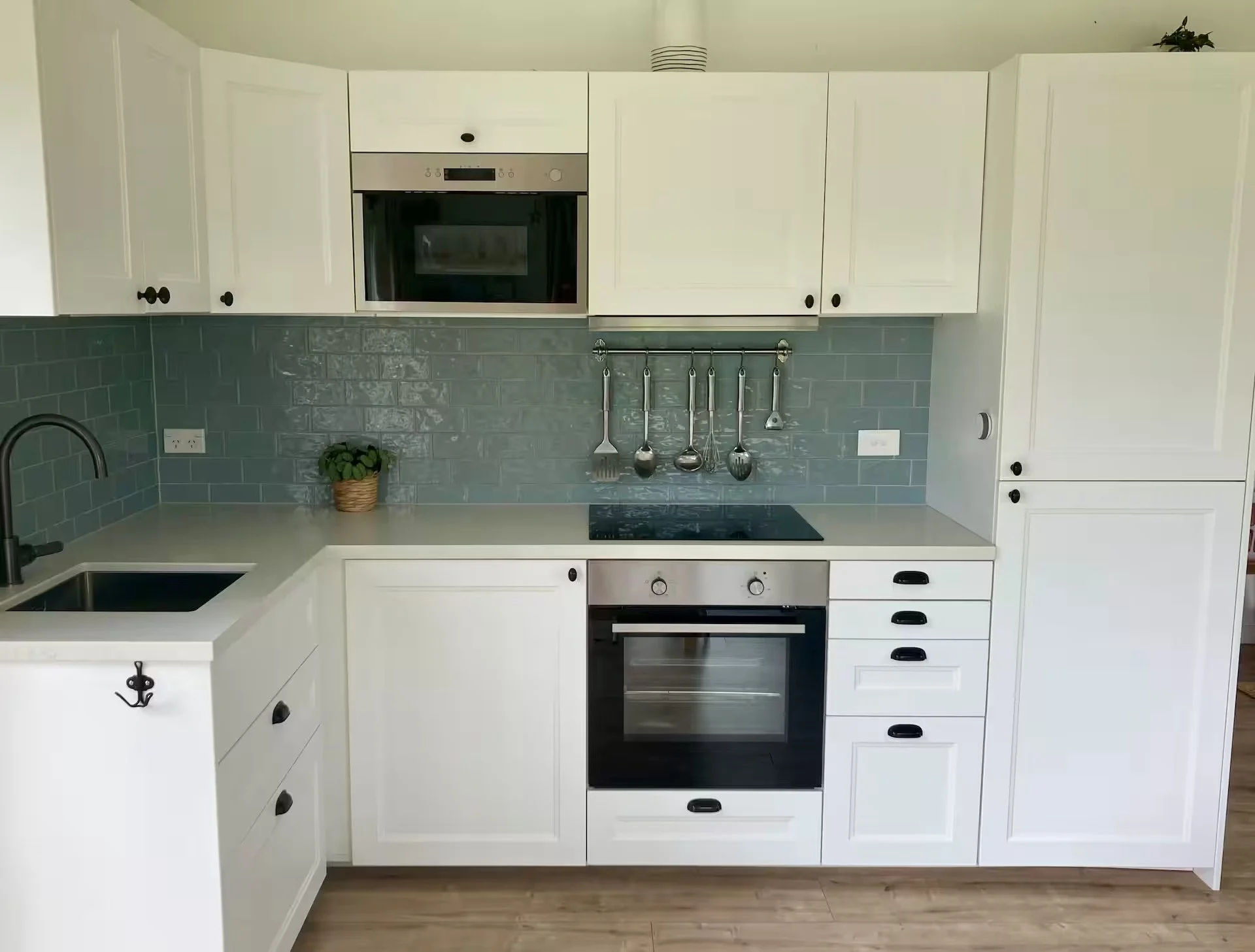Modern kitchen with white cabinets, a stainless steel oven, a black stovetop, and teal subway tile backsplash. A small potted plant is on the counter, and kitchen utensils hang from a rail.