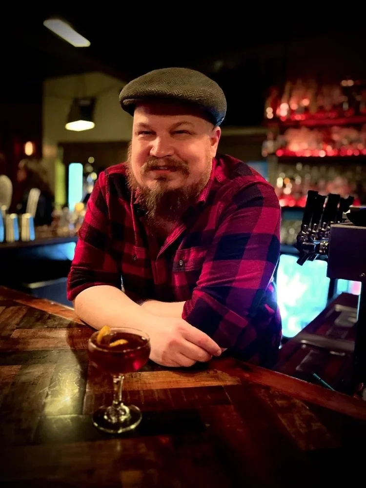 A man with a beard and mustache wearing a gray flat cap and red plaid shirt, sitting at a wooden bar counter with a cocktail in front of him, in a dimly lit bar or pub.