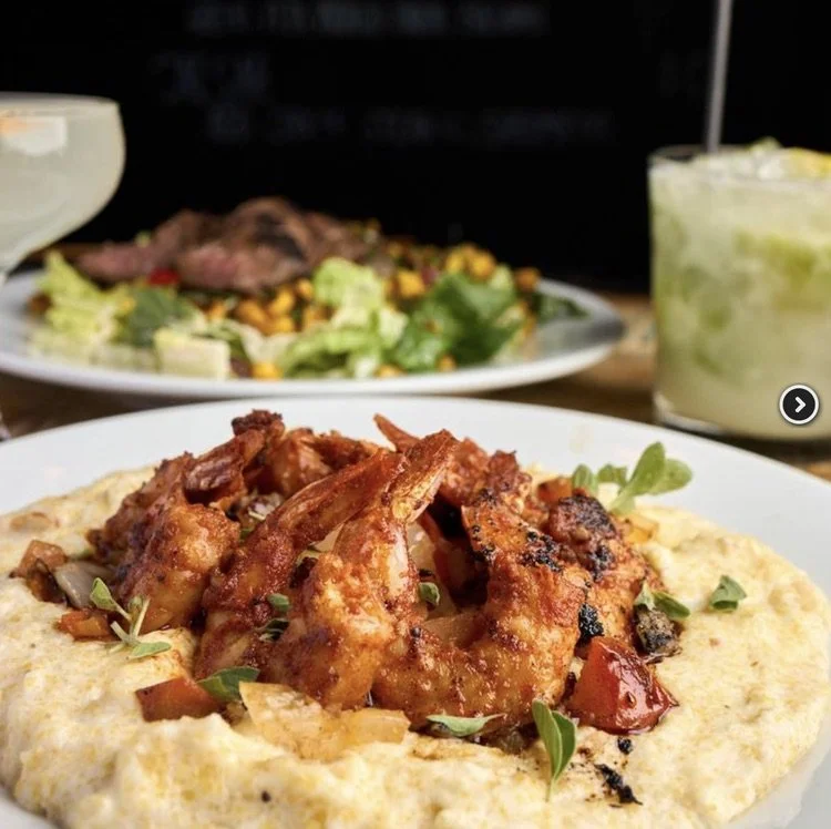 Plate of grilled shrimp with mixed vegetables and a side of mashed potatoes, with a salad and a glass of lime drink in the background.