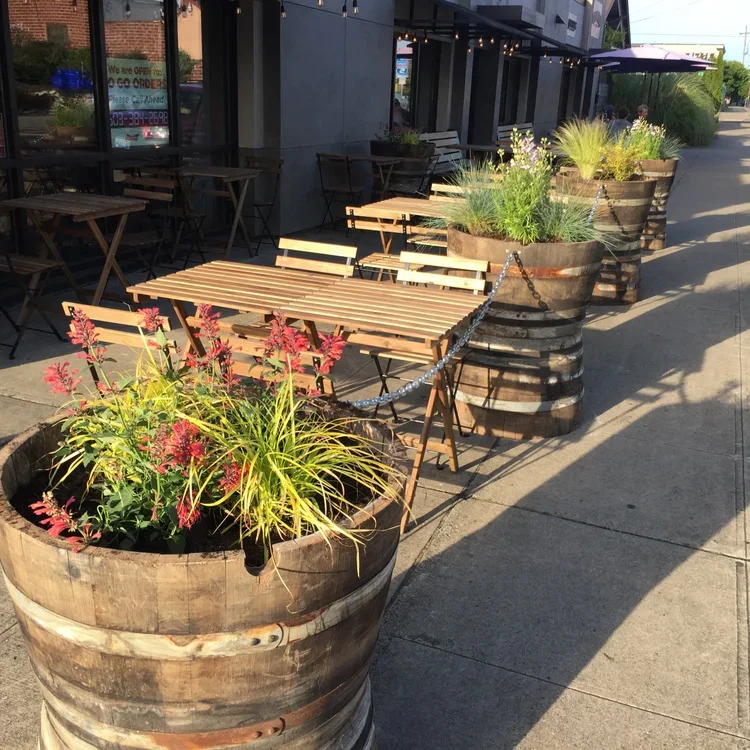 Outdoor sidewalk seating area with wooden tables and chairs, large wooden barrel planters filled with various green plants and flowers, chain barrier separating seating from the sidewalk, sunlight casting shadows.
