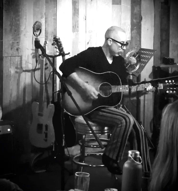 A person playing an acoustic guitar on a small stage in a music venue with wooden walls, wearing glasses and striped pants, and an audience member visible in the foreground.