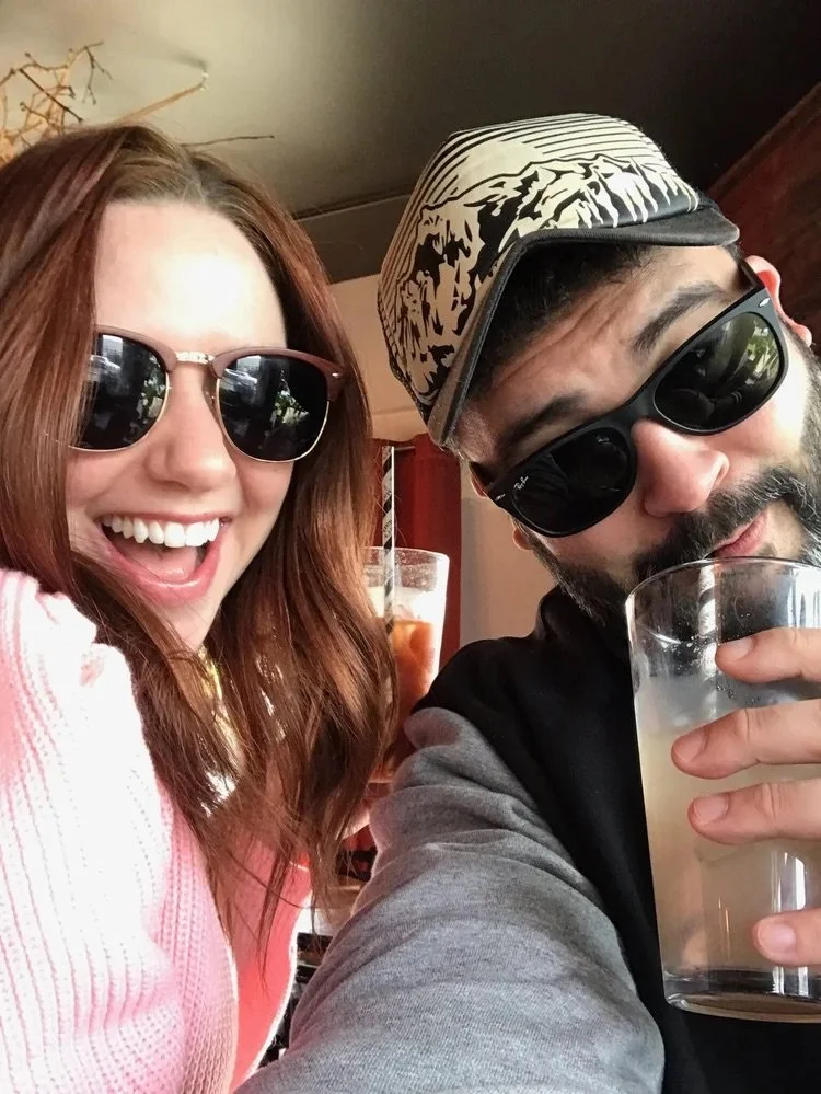 A woman with red hair wearing sunglasses and a pink top, smiling broadly. A man with a beard, sunglasses, wearing a patterned cap, holding a drink, taking a selfie with her.