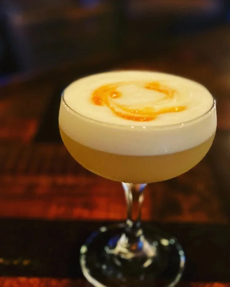 Cocktail with creamy white foam and a caramel swirl garnish served in a coupe glass on a dark wooden surface.