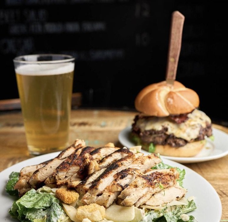 A plate of grilled chicken salad with lettuce and croutons, a cheeseburger with lettuce and bacon, and a glass of beer on a wooden table.