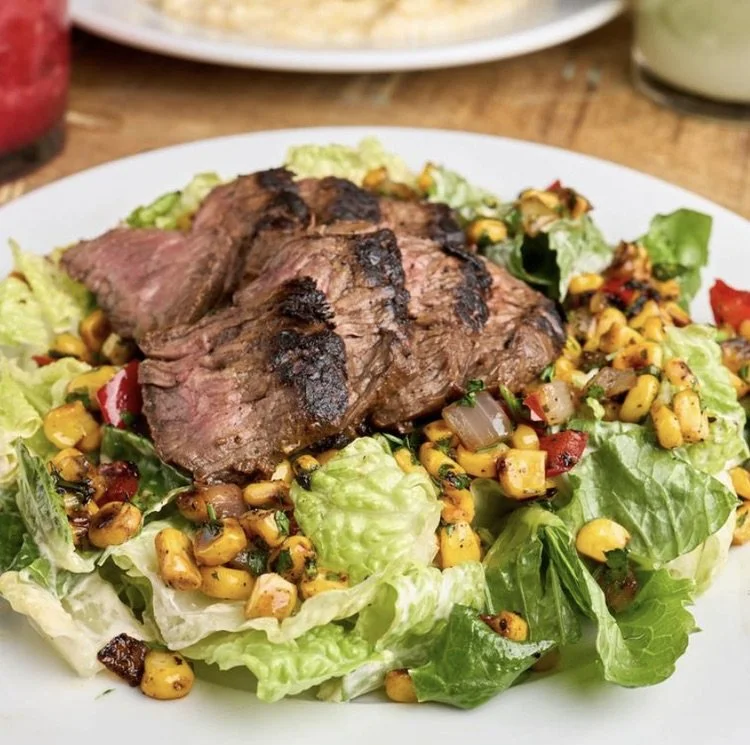 Grilled steak on a bed of mixed salad greens with corn, chopped vegetables, and herbs.