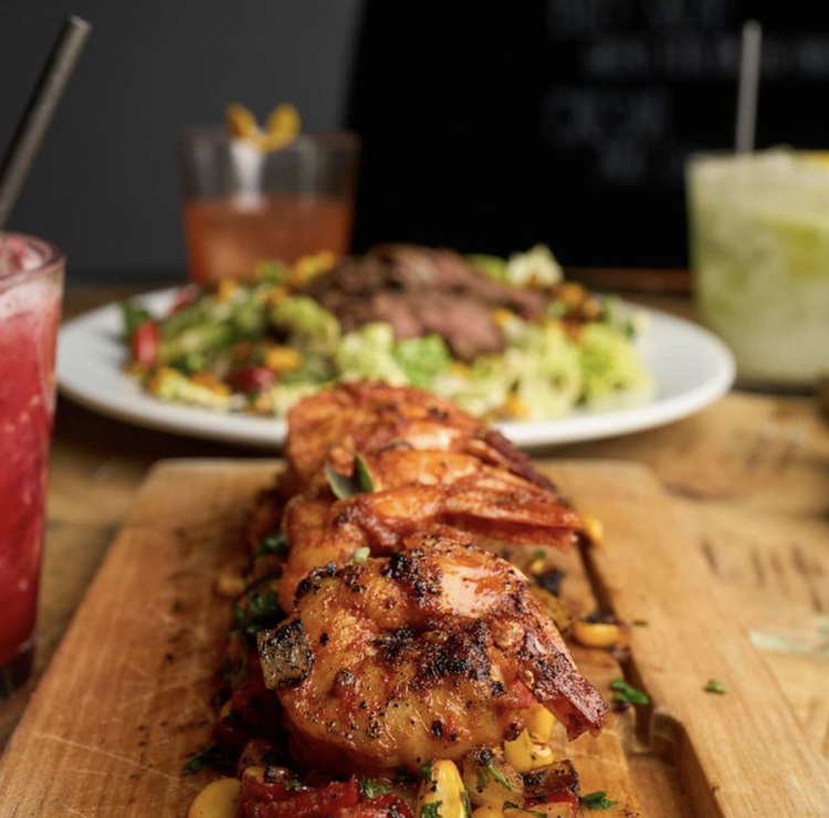 Close-up of grilled chicken skewers with vegetables on a wooden board, with a salad, drinks, and other dishes in the background.