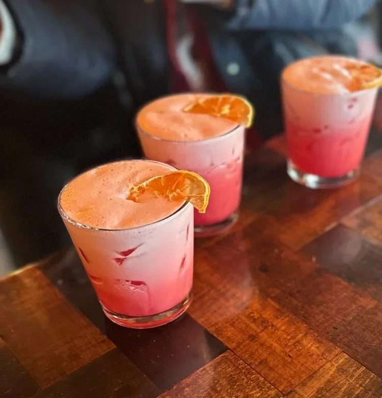 Three pinkish cocktails with orange slices garnishing, served in short, clear glasses on a wooden bar counter.