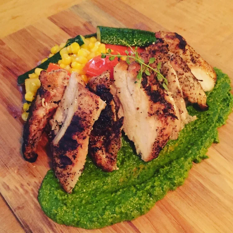 Grilled chicken breast slices served on a bed of green sauce, with side of corn, cucumber, and tomato on a wooden platter.