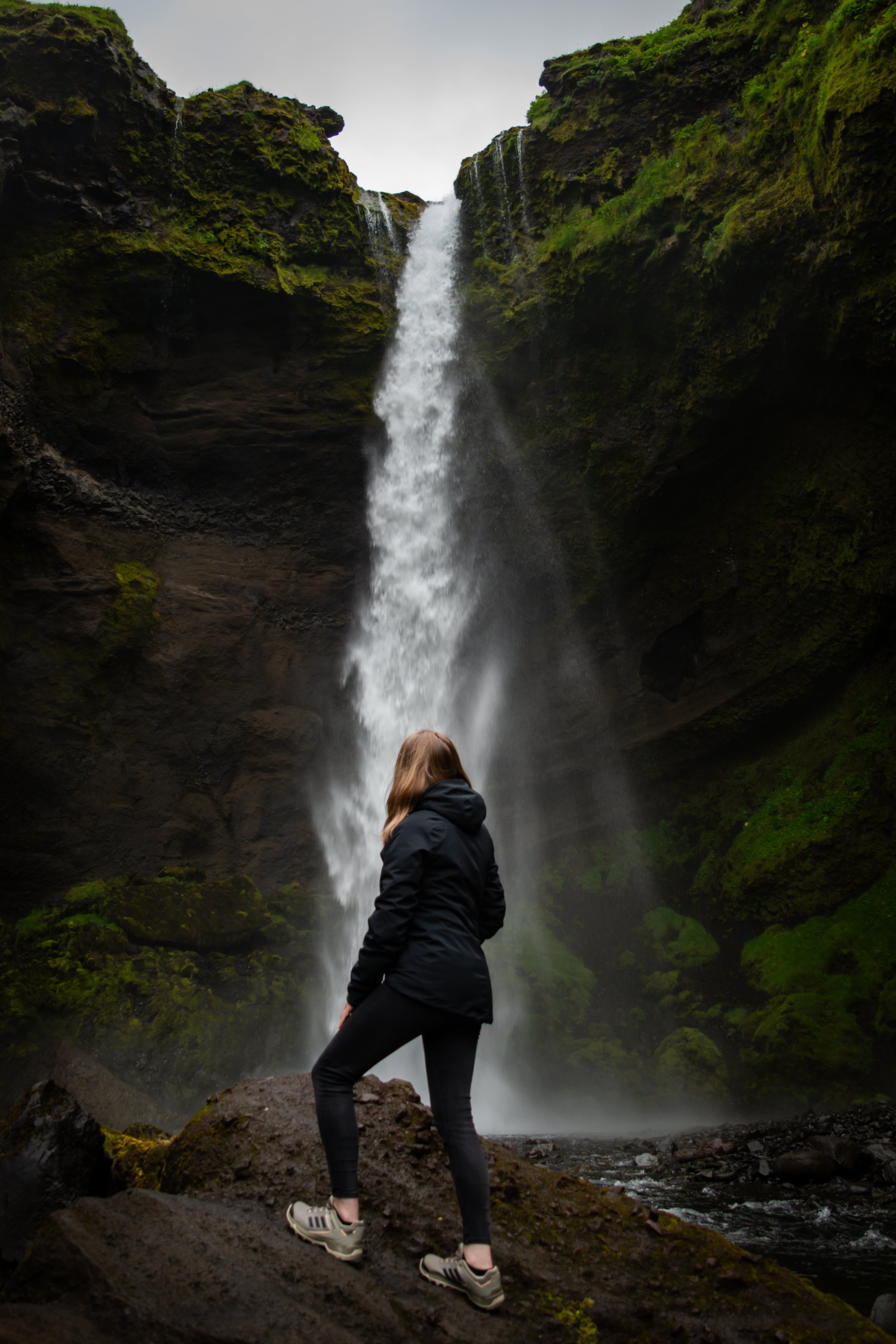 A person in dark clothing and beige sneakers stands on a rock in front of a tall waterfall cascading down moss-covered cliffs, with the person facing the waterfall.