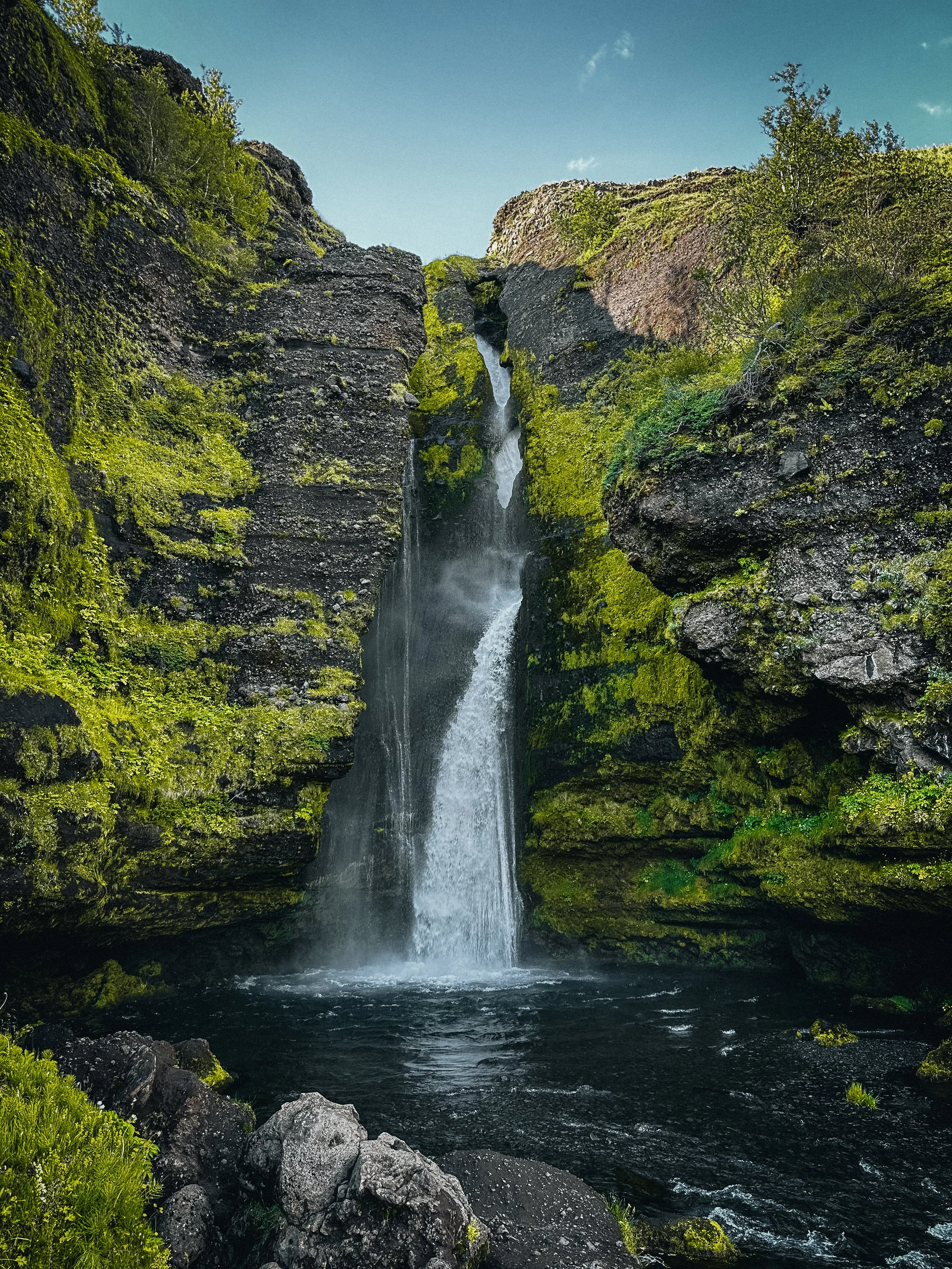 A waterfall flowing down moss-covered rocks into a dark pool of water surrounded by green vegetation under a blue sky.