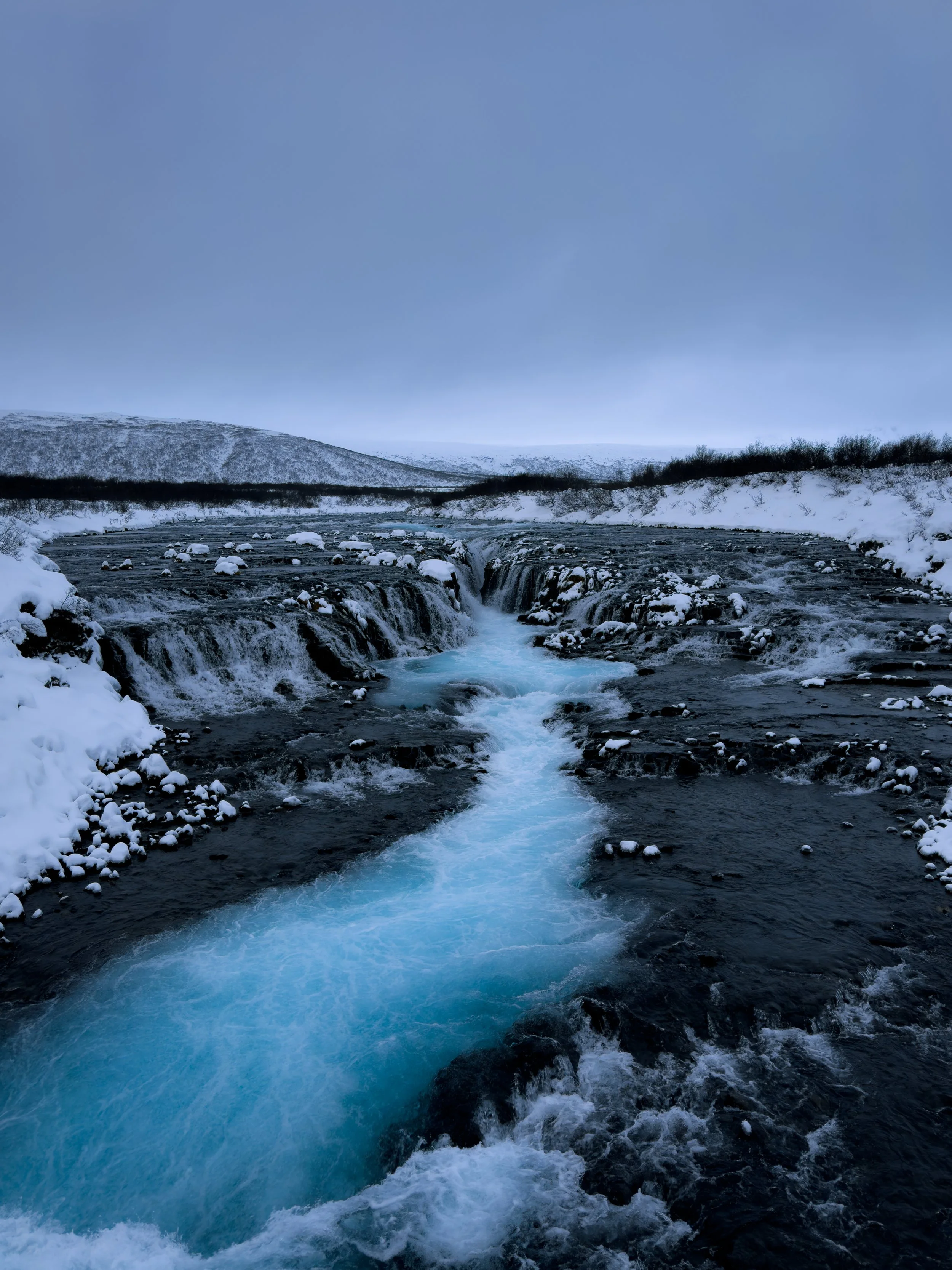 A river flowing through a snow-covered landscape with small waterfalls, surrounded by snow and trees under a cloudy sky.