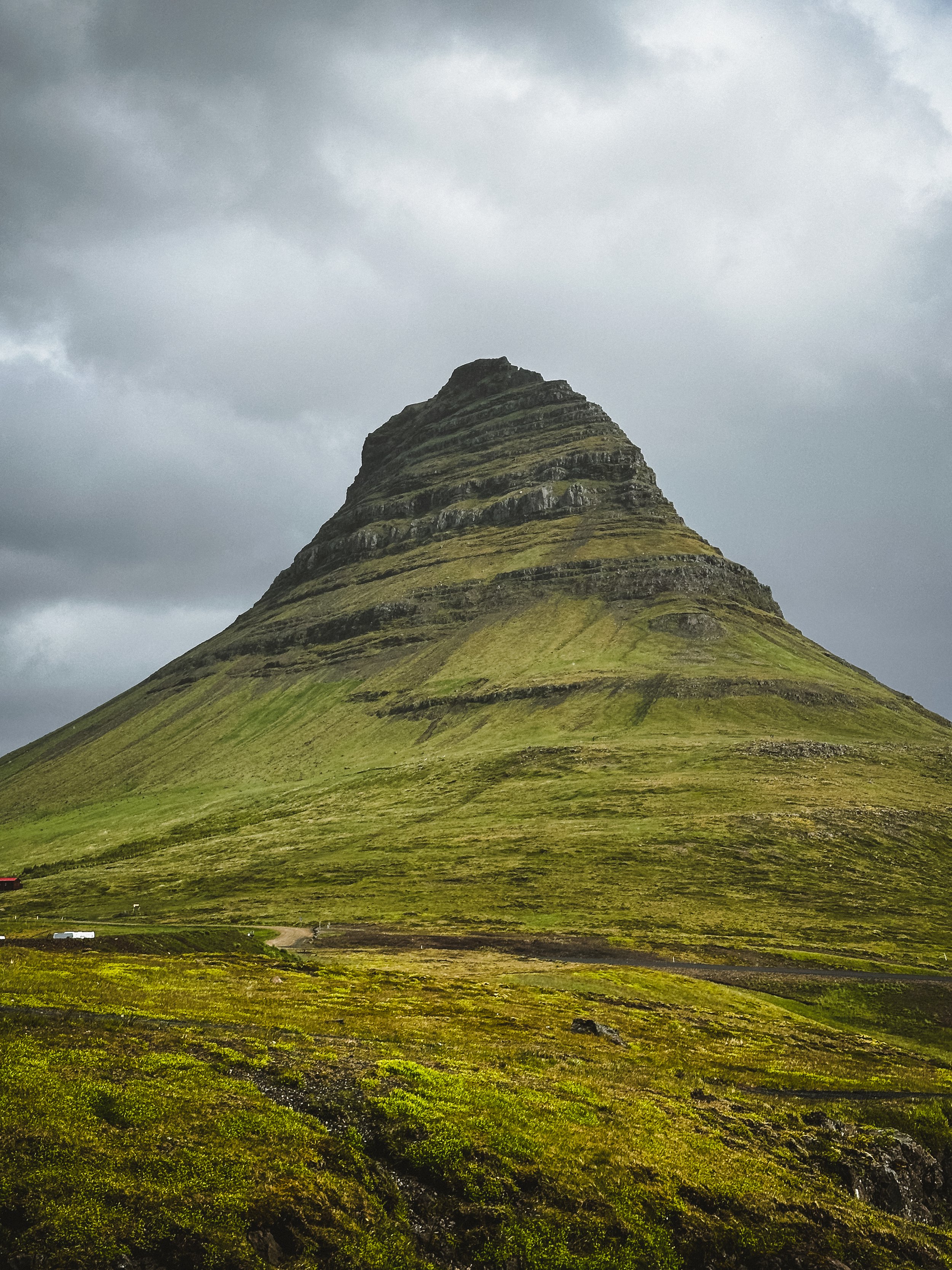 A green, layered mountain with a conical shape under a cloudy sky.