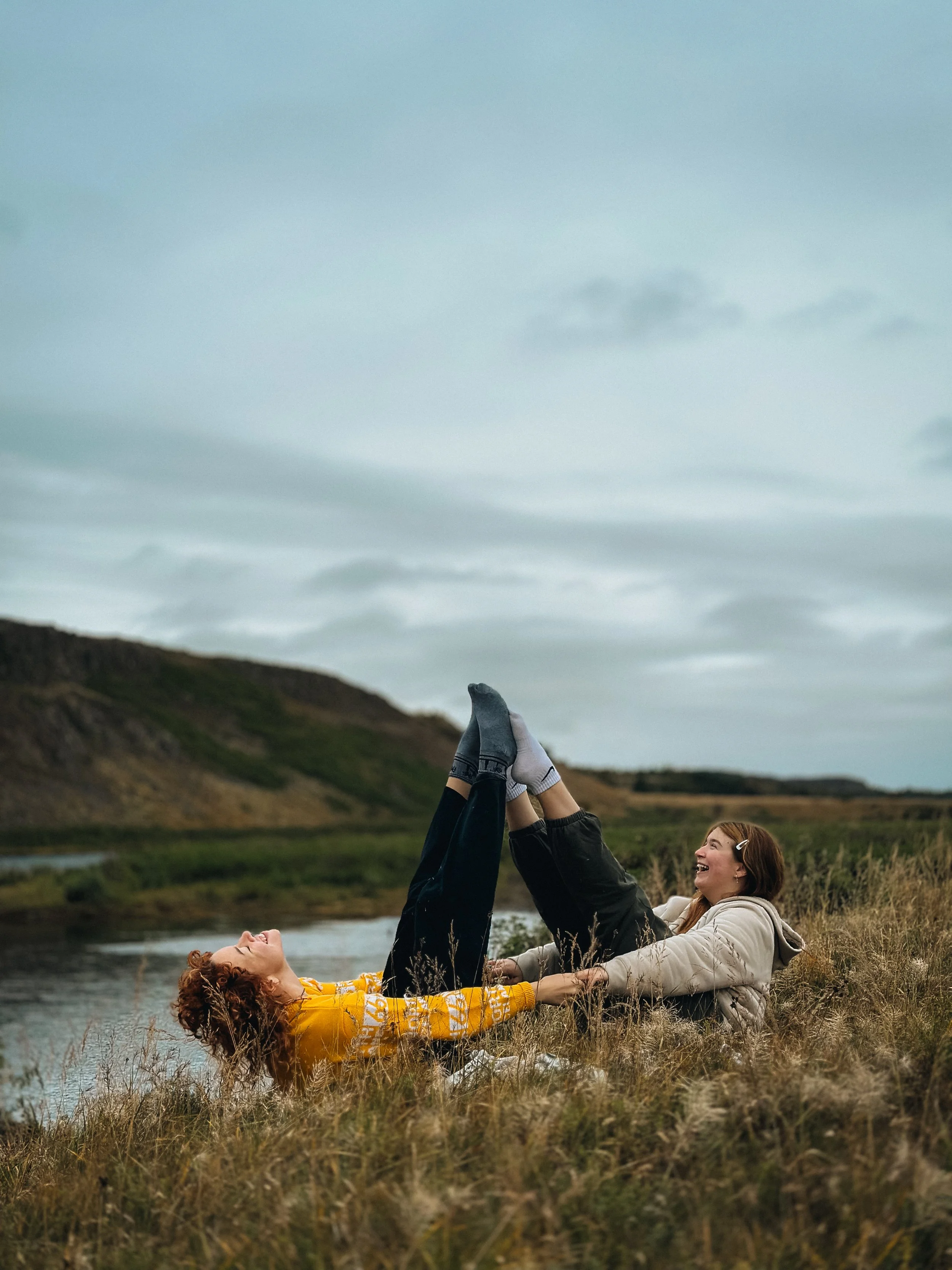 Two women are lying on the grass near a river, holding hands and raising their legs in the air, laughing and enjoying the outdoor scenery with hills and cloudy sky in the background.