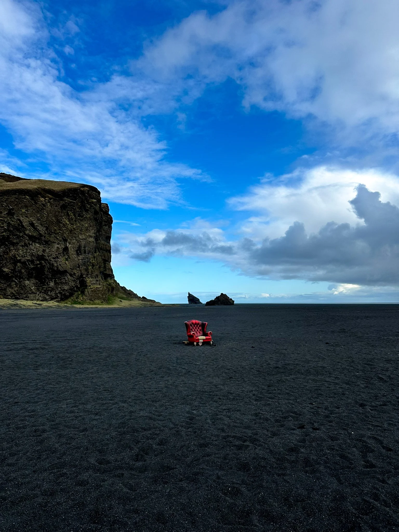 A red velvet armchair on black sand beach with cliffs and ocean in the background, under a partly cloudy sky.