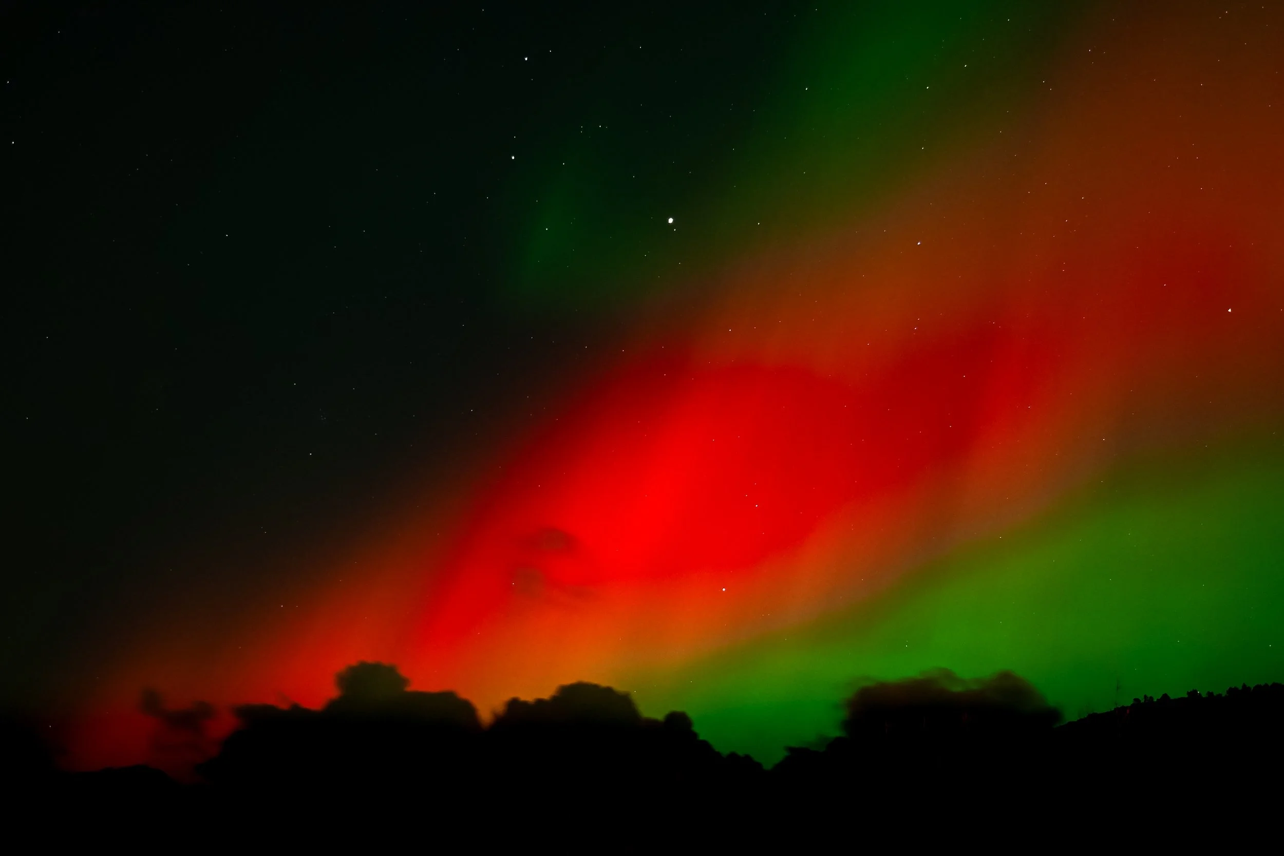 Northern lights displaying green and red hues above a silhouette of a mountain landscape at night.