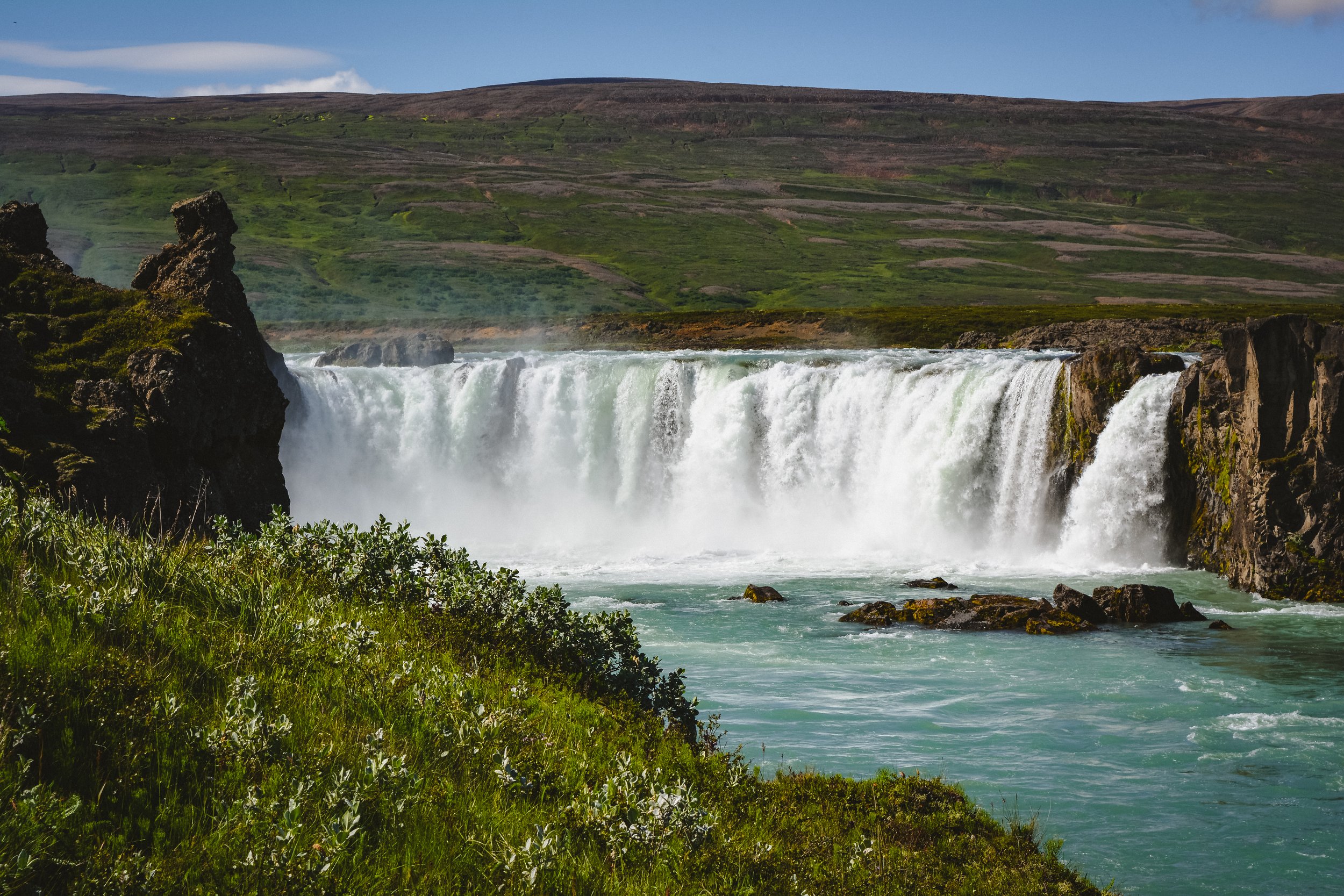 A large waterfall cascading over a rocky cliff into a wide river, surrounded by green grass and bushes, with a hill covered in vegetation in the background under a partly cloudy sky.