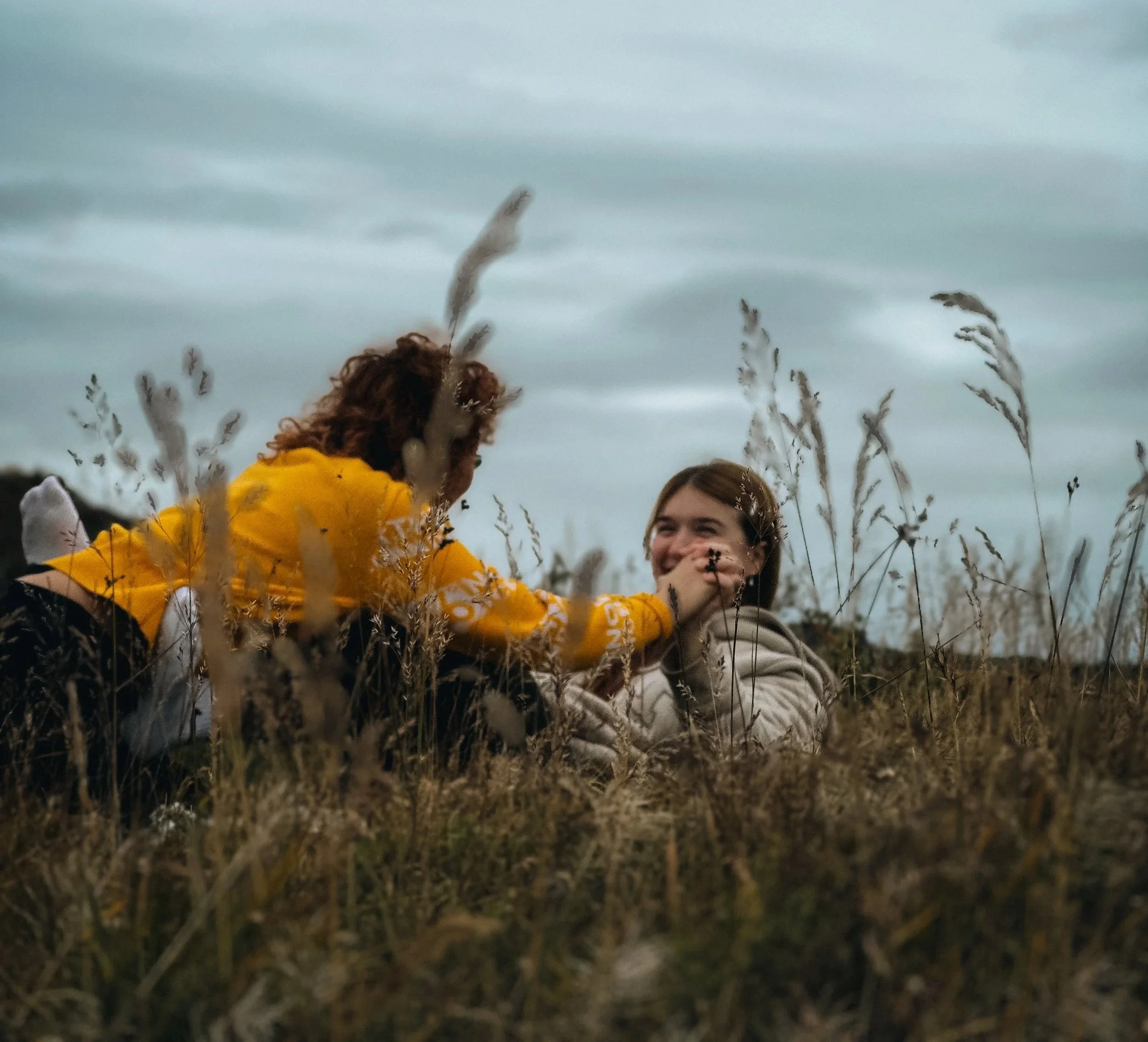 Two young women lying on the grass in a field with tall, dry grass, one wearing a yellow jacket and the other a beige jacket, smiling and playing with each other's faces.