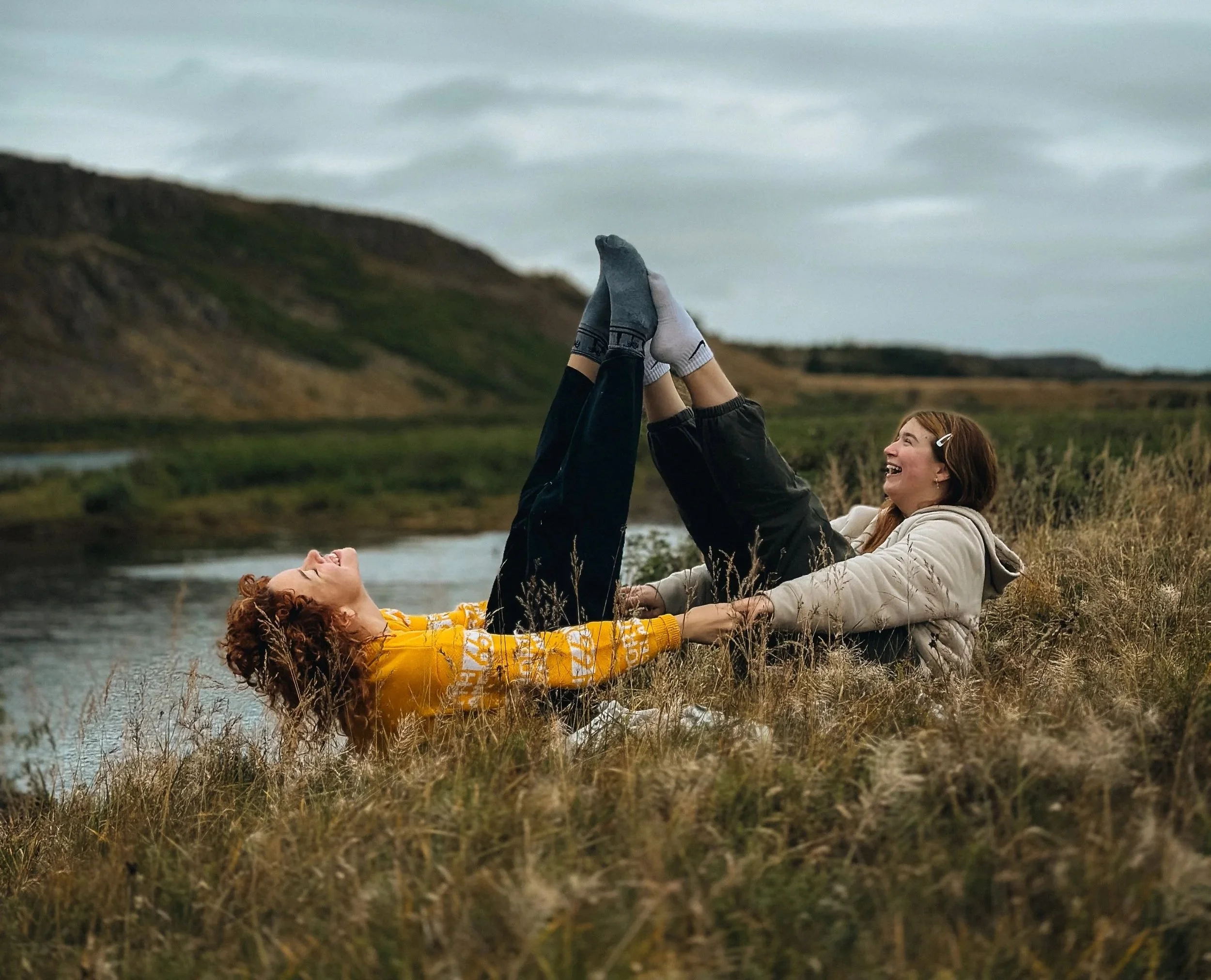 Two women playing and laughing outdoors near a river with grassy fields and mountains in the background
