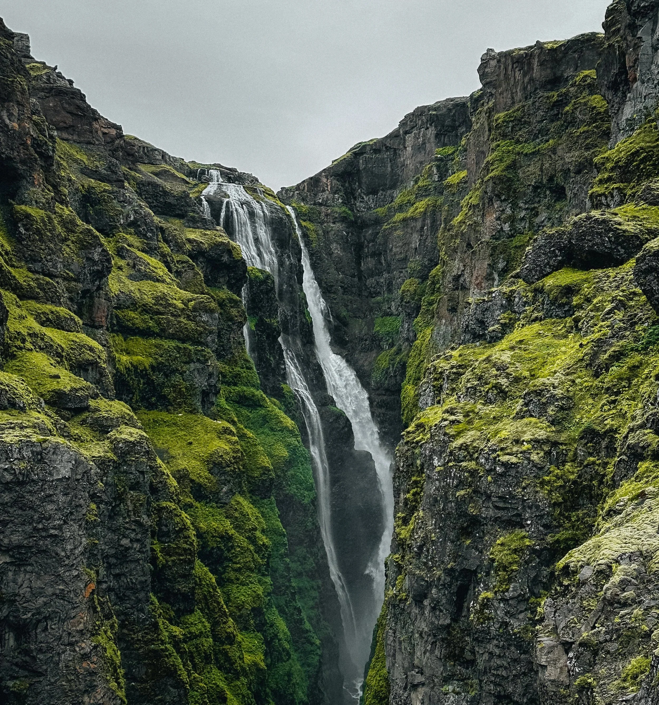 A tall waterfall cascading down moss-covered rocky cliffs in a narrow ravine, with gray overcast sky.