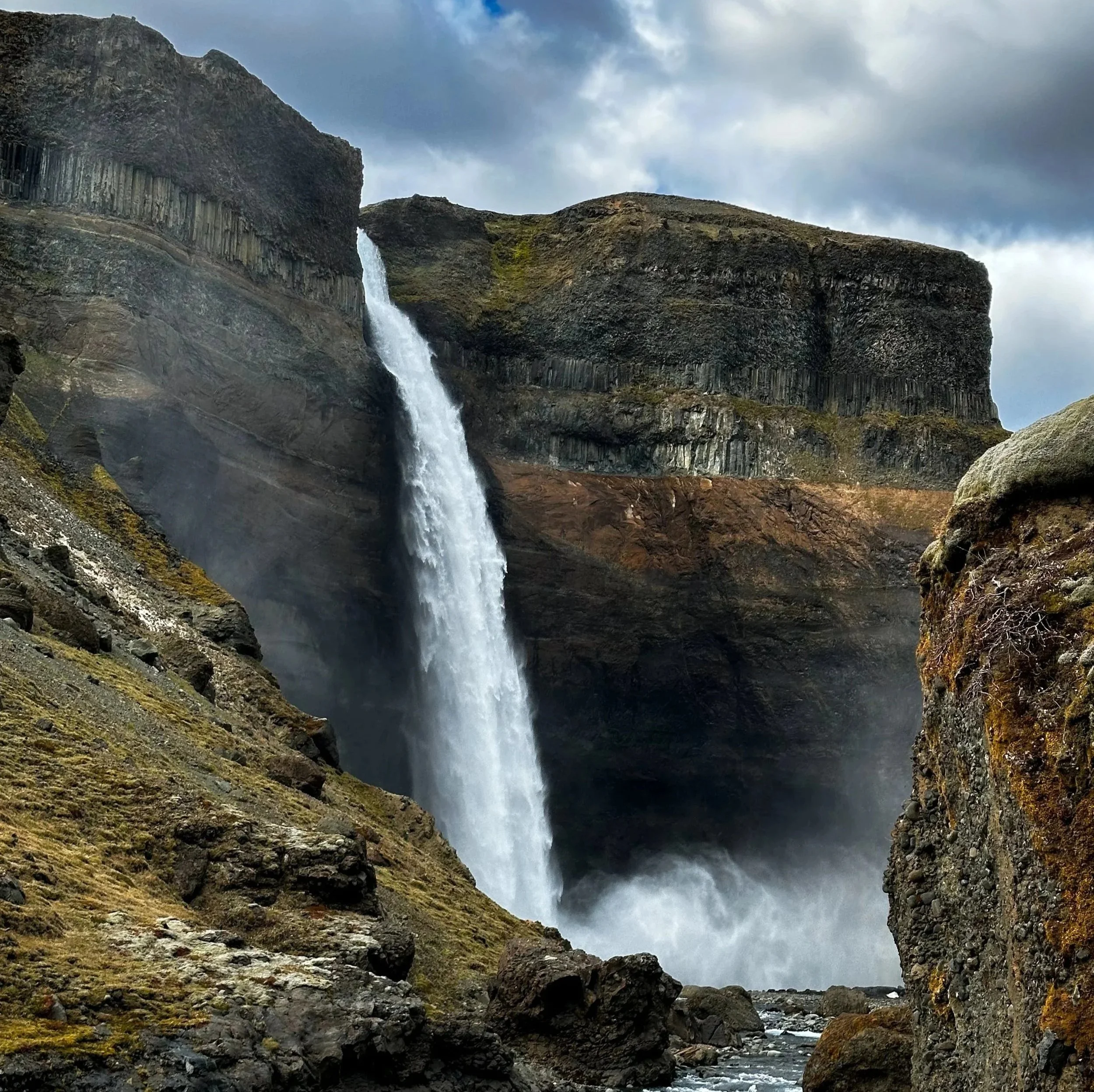 A waterfall cascading down a rocky cliff into a river below, surrounded by rugged terrain and moss-covered rocks, with overcast skies.