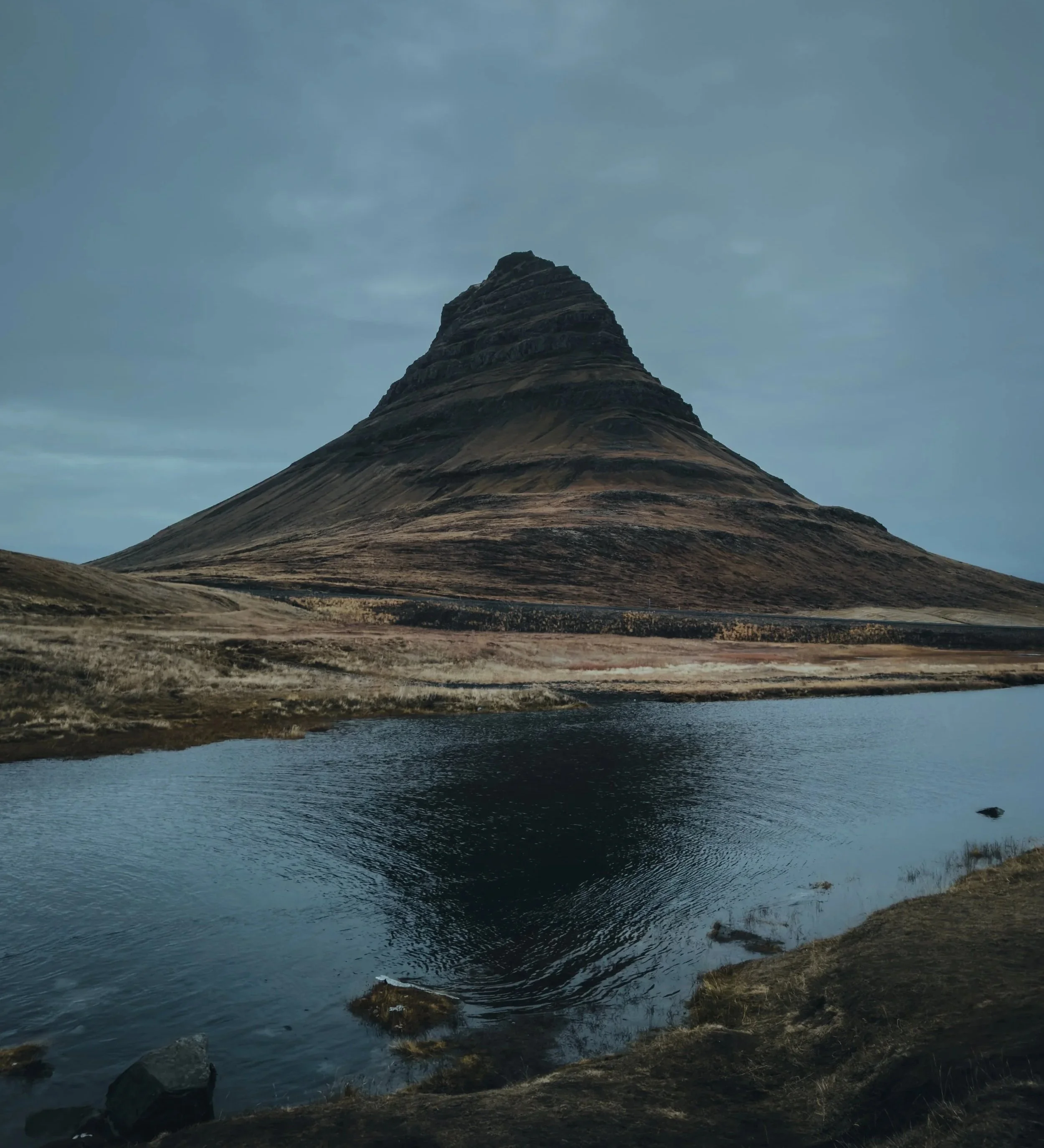 A mountain with a conical shape reflecting in a body of water, surrounded by a grassy landscape under a cloudy sky.