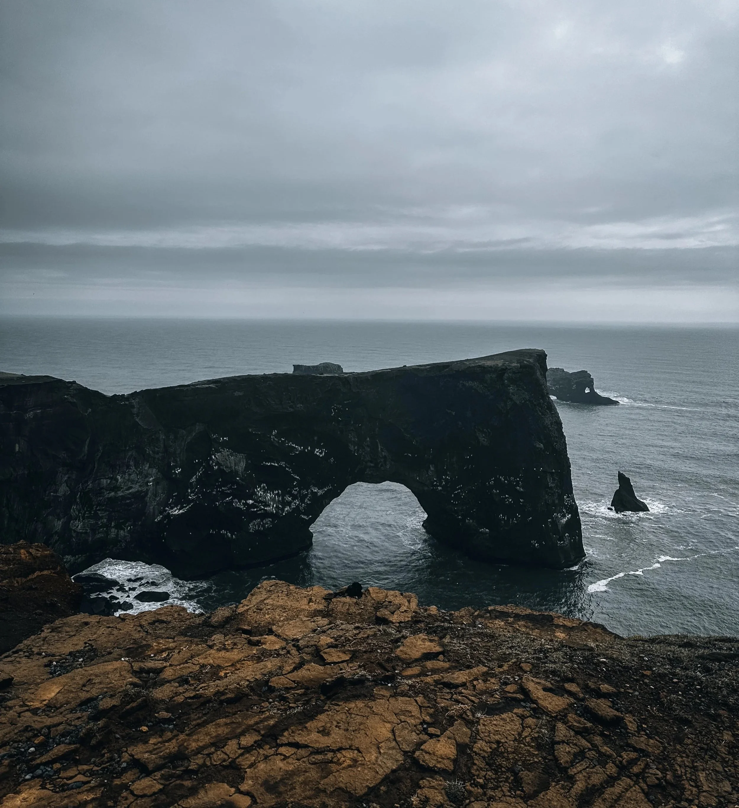 A dark, rocky coastal landscape with a natural rock arch formation over the water under a cloudy sky.