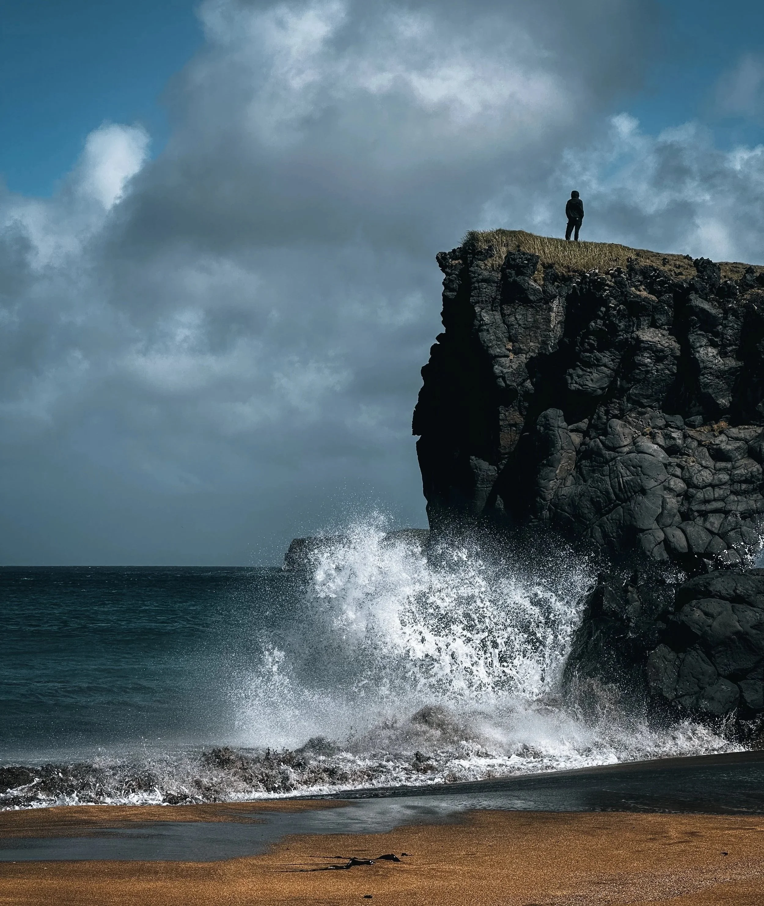 A person standing on a grassy cliff overlooking the ocean as waves crash against the rocks below under a cloudy sky.