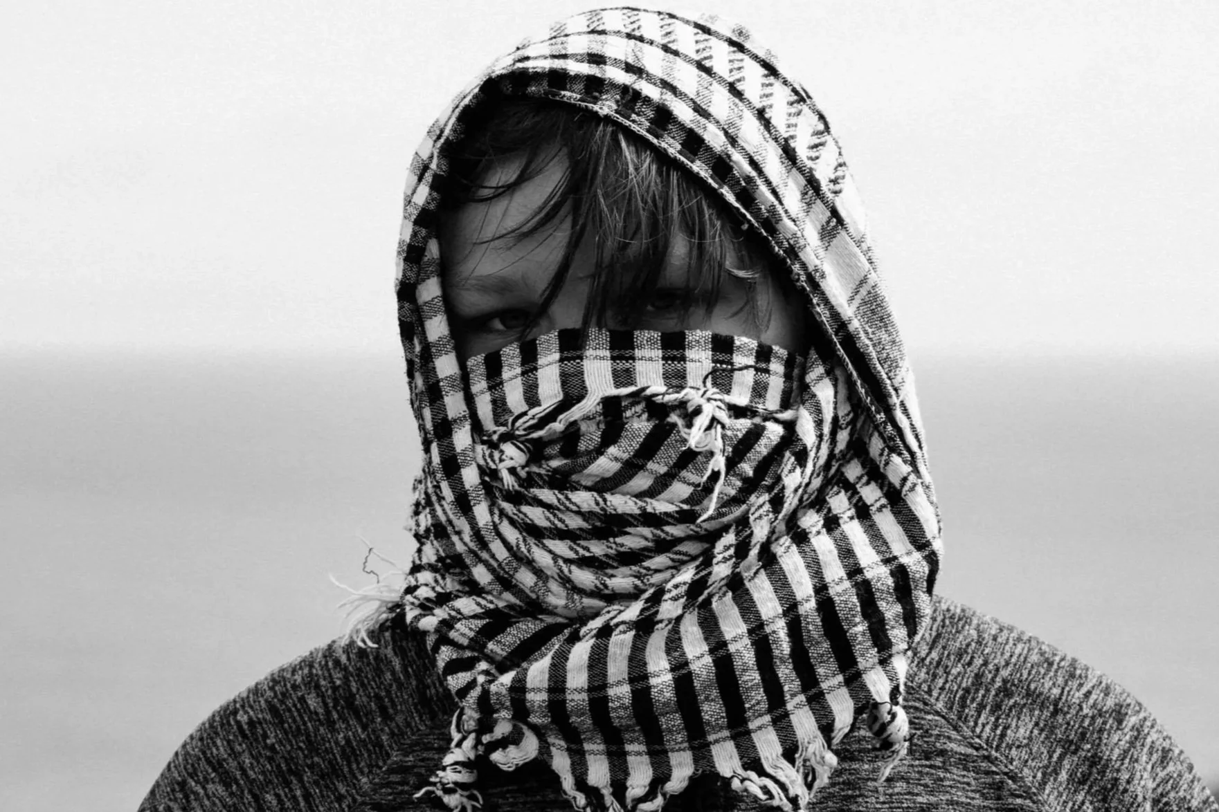 A person with a checkered headscarf covering their face, revealing only one eye, against a plain background.