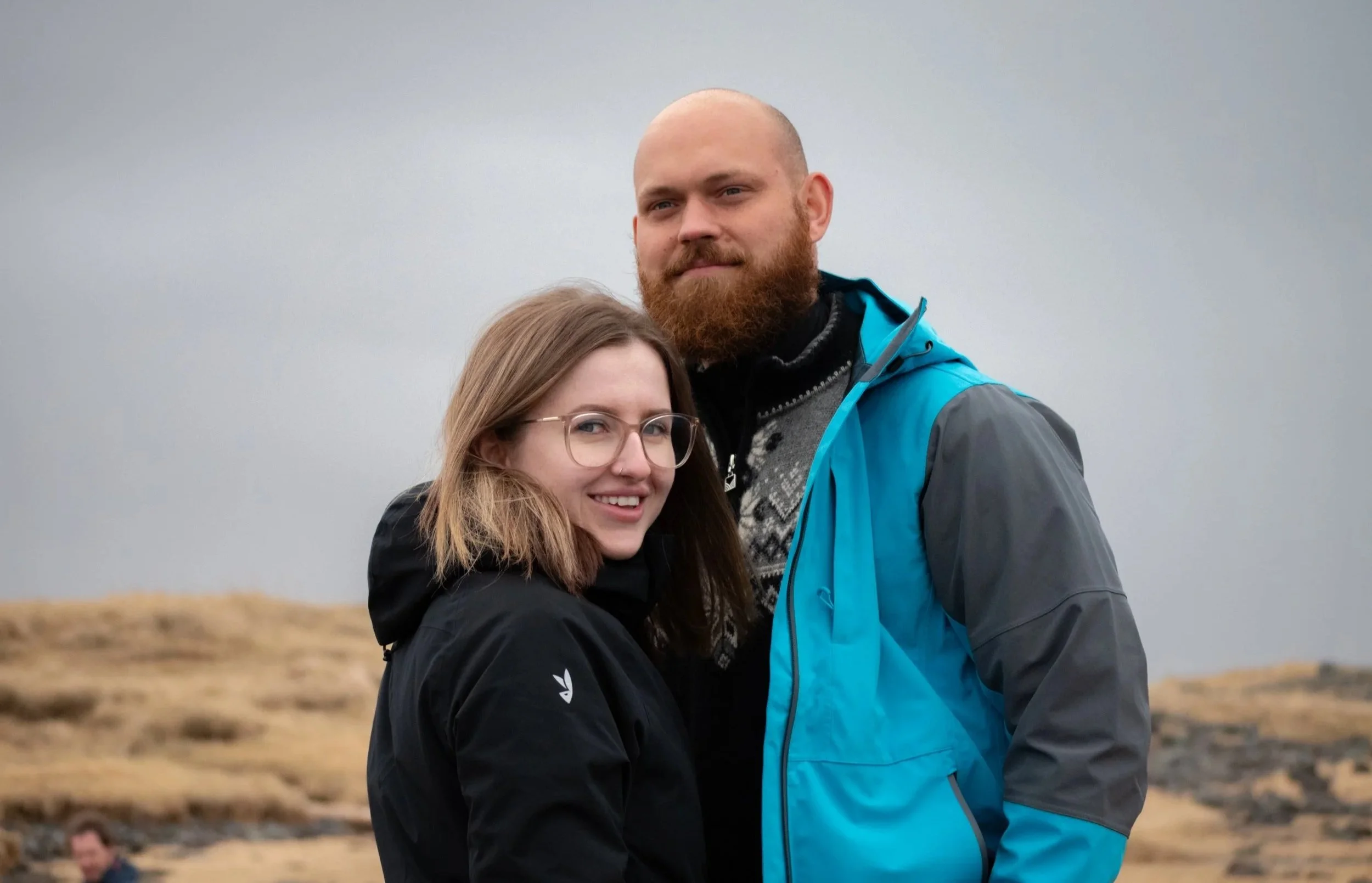A man with a beard and a woman with glasses are standing outdoors on a cloudy day, smiling and posing for the photograph.