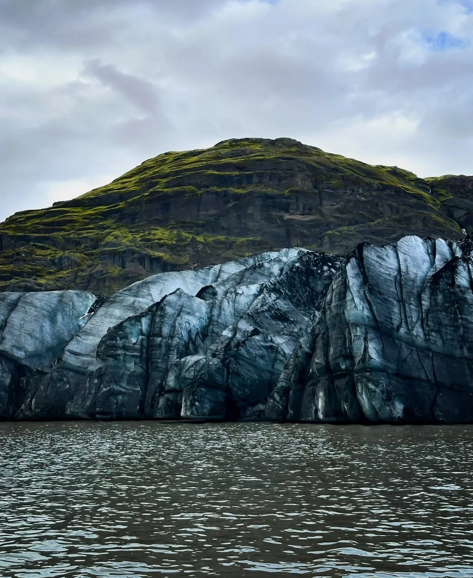 A large glacier with black streaks, sitting at the base of a green, moss-covered mountain, with water in the foreground.
