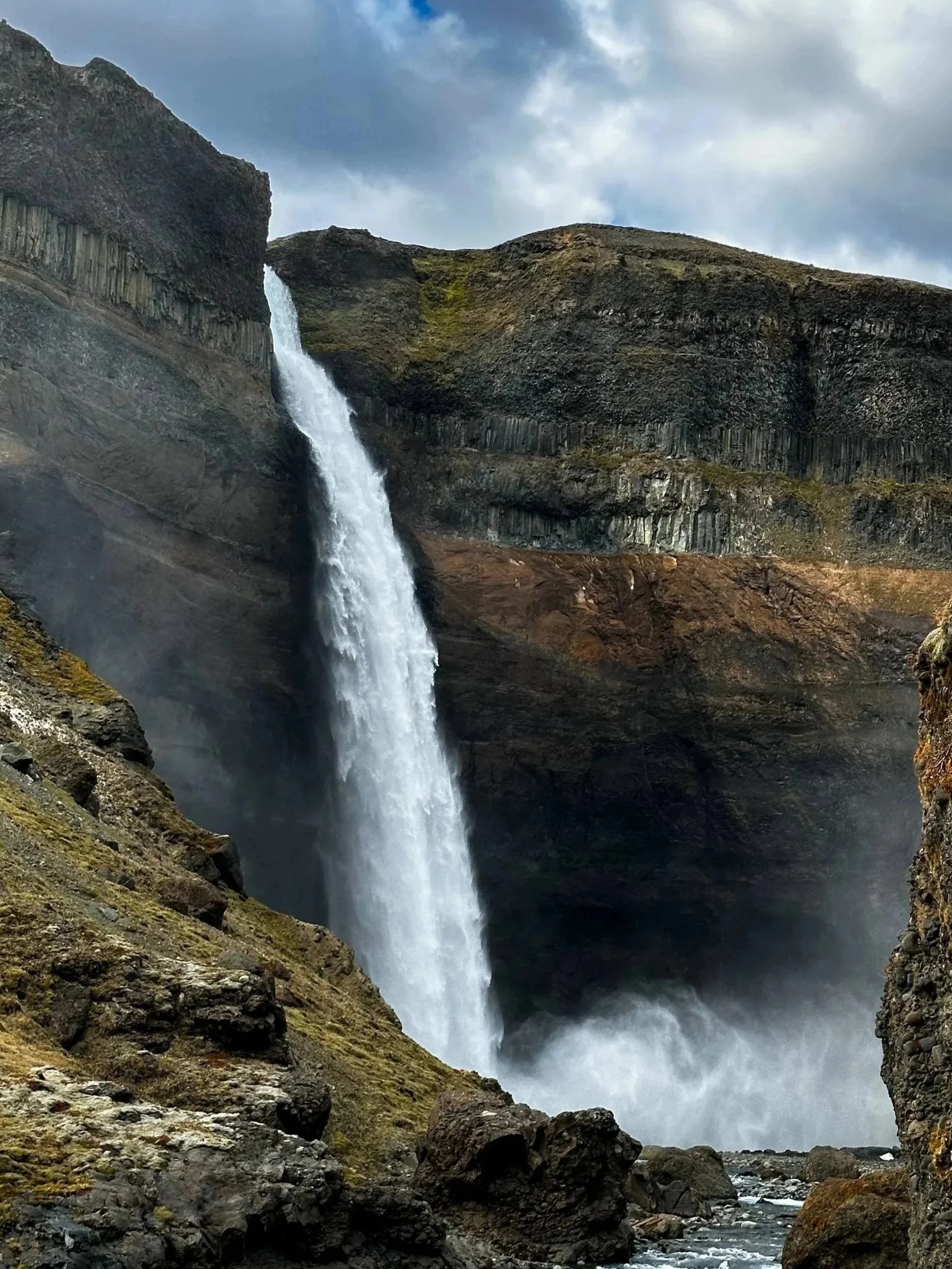 A waterfall cascading down a rocky cliff into a river below, surrounded by rugged terrain and moss-covered rocks, with overcast skies.