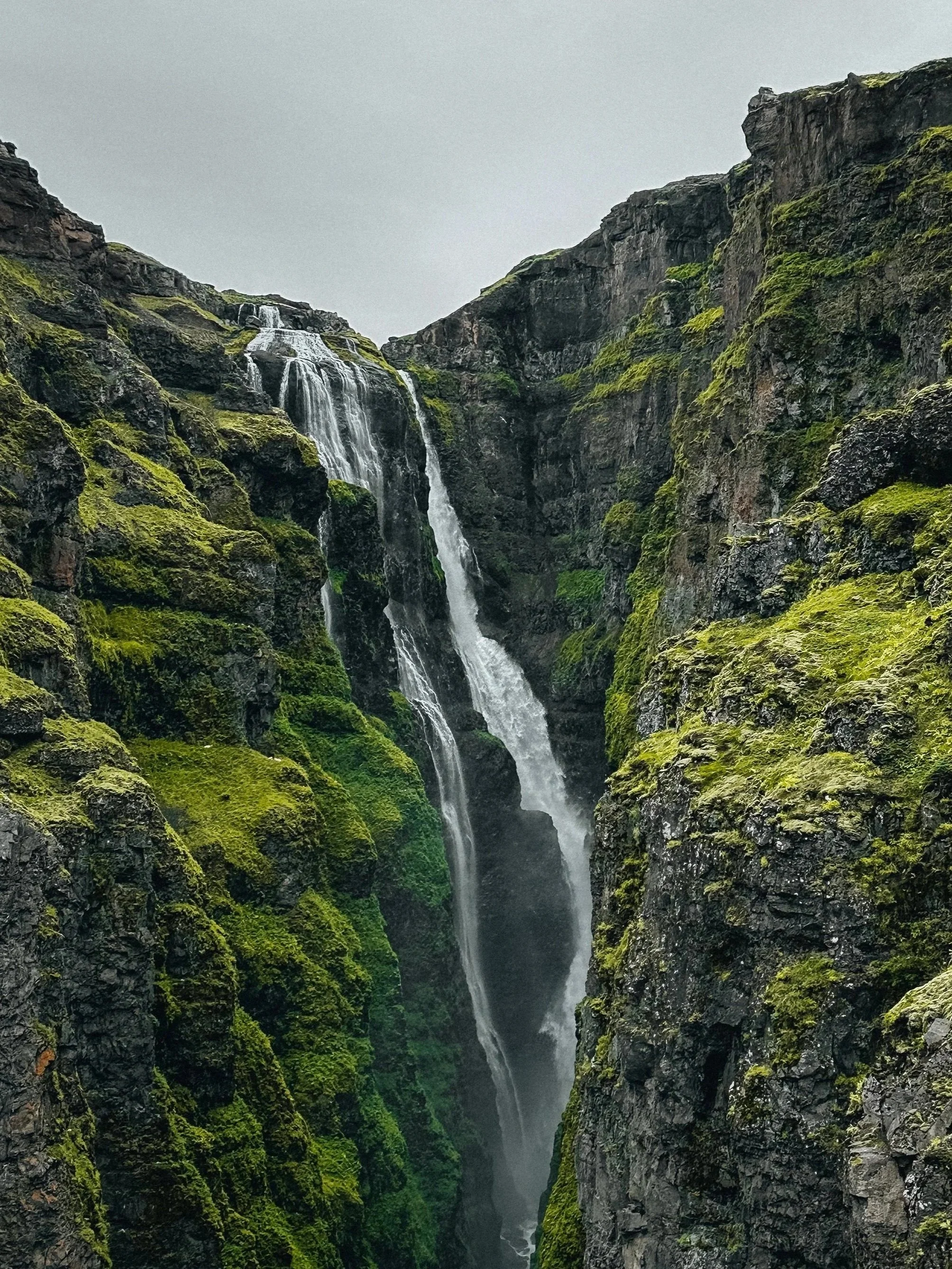 A tall waterfall cascading down moss-covered rocky cliffs in a narrow ravine, with gray overcast sky.